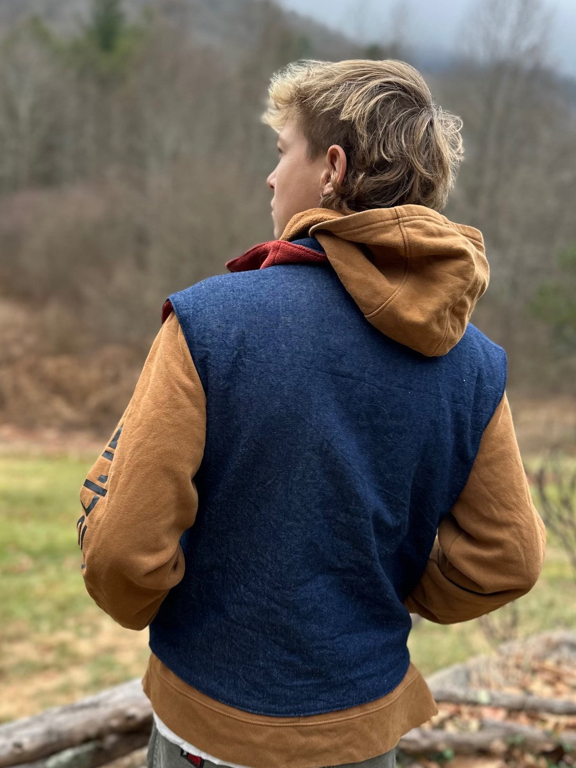Person wearing a blue vest over a brown hoodie with a forest background. Back to camera