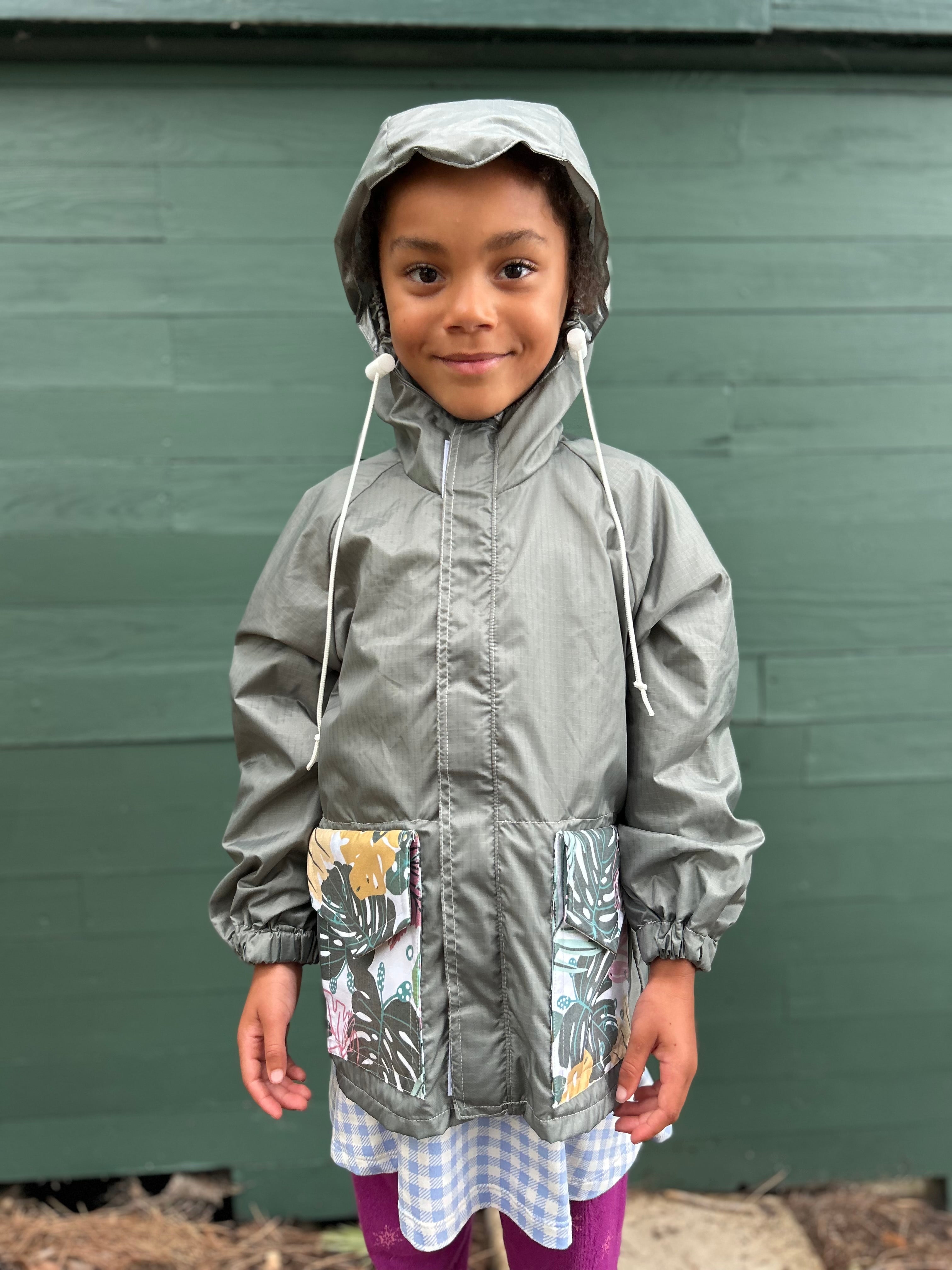 A young child wearing a gray hooded raincoat with colorful tropical floral print pockets, standing in front of a dark green wooden wall.
