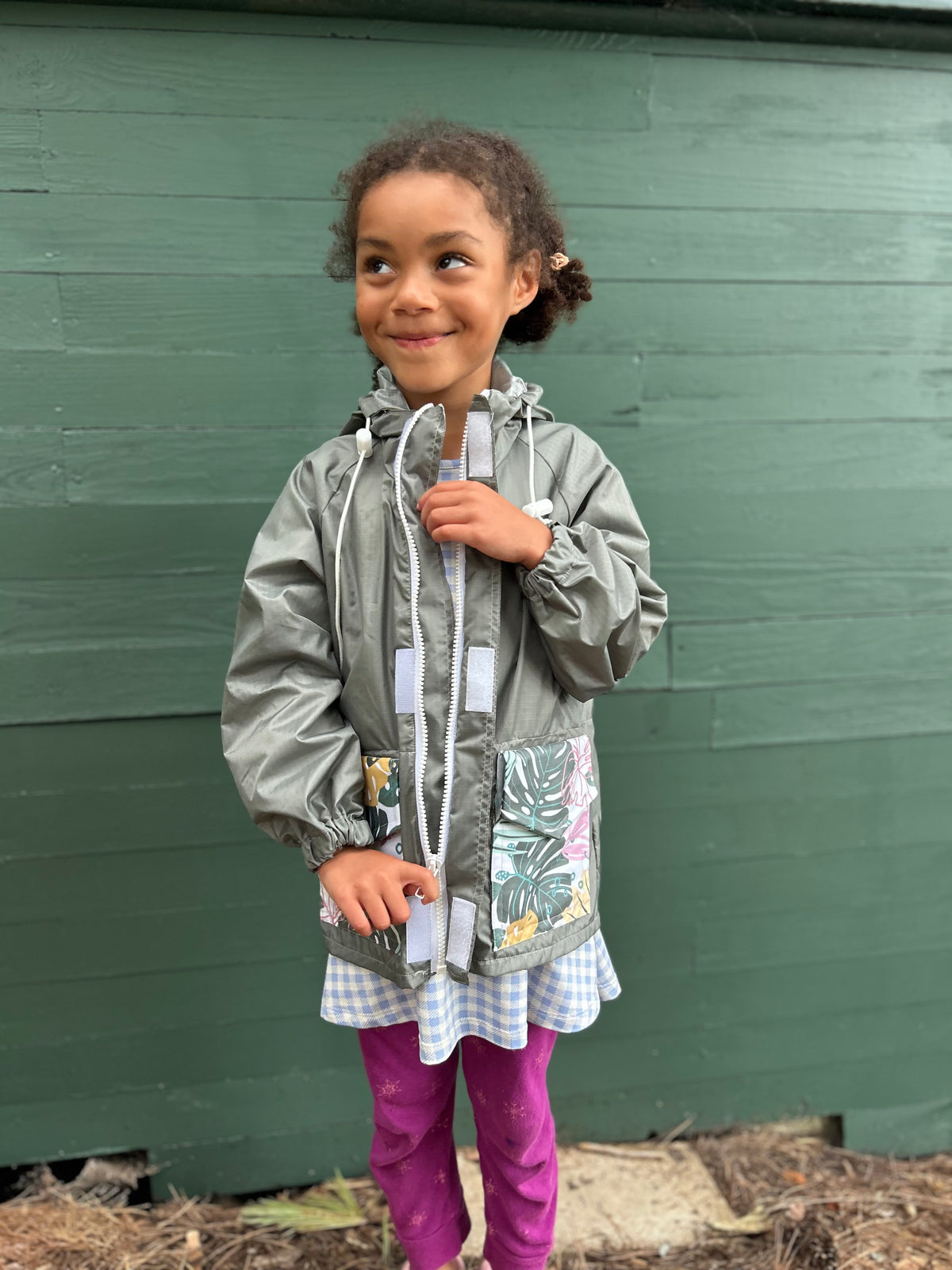 A young girl with curly brown hair stands in front of a dark green wooden wall, wearing a silver-gray hooded raincoat with a colorful tropical leaf print pocket, a blue and white gingham dress, and magenta leggings.