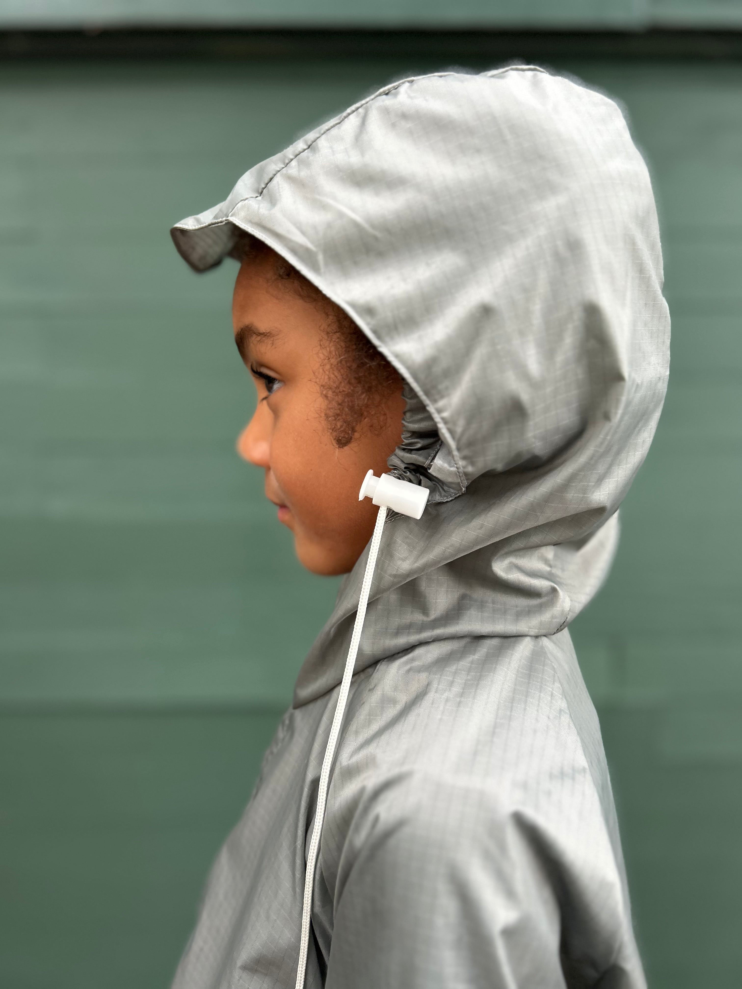 A child in profile wearing a light gray hooded raincoat with a white drawstring, standing against a muted green wall.