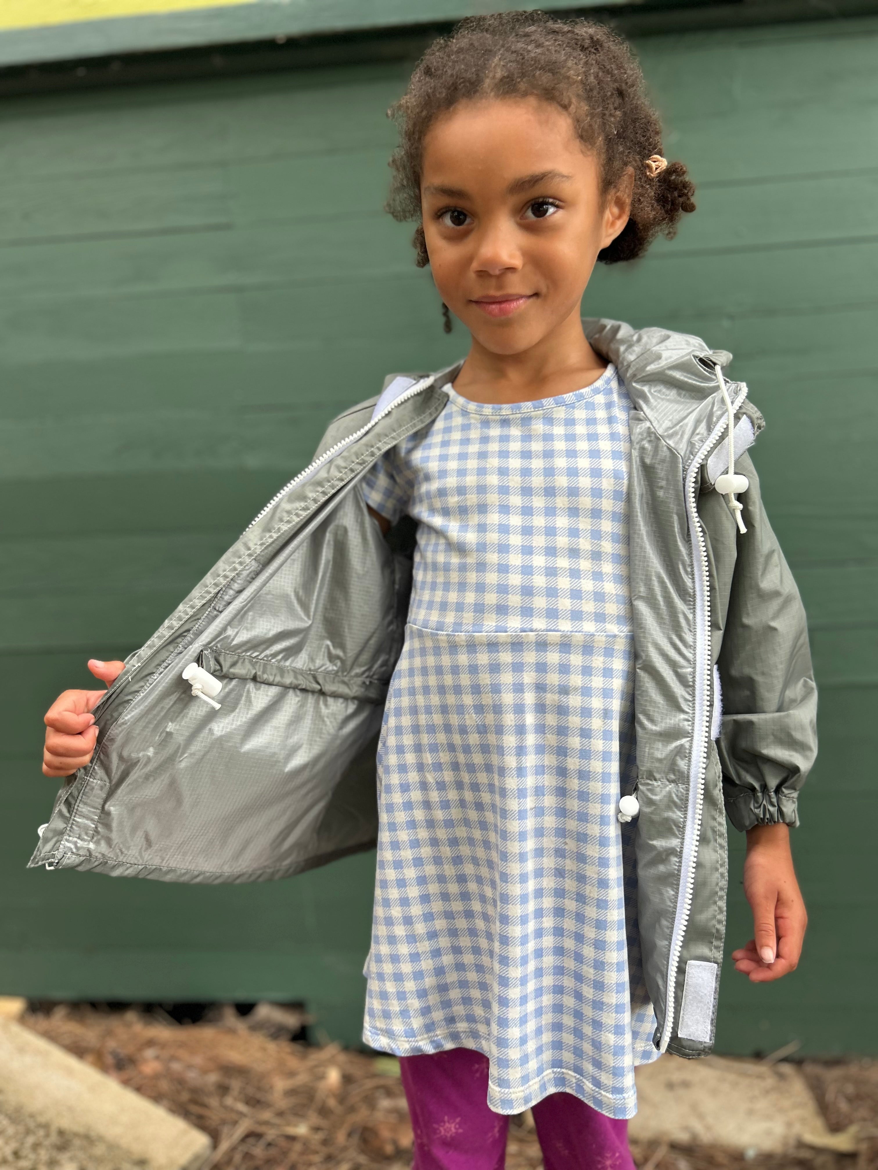 A young girl wearing a light blue and white gingham dress under an open silver hooded jacket, standing in front of a dark green wooden wall.