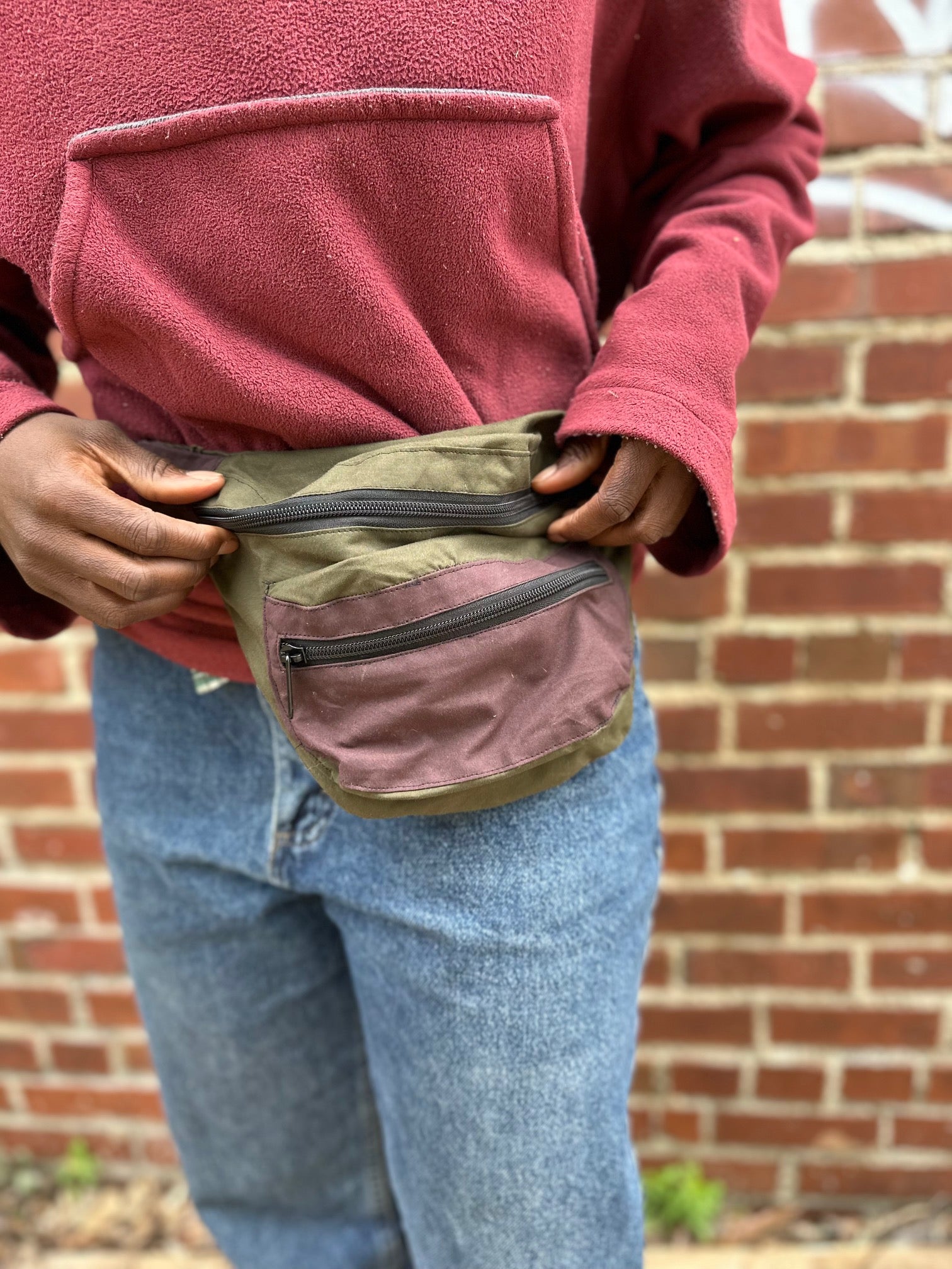 Person wearing a pink fleece jacket and blue jeans with a green waist bag against a brick wall.