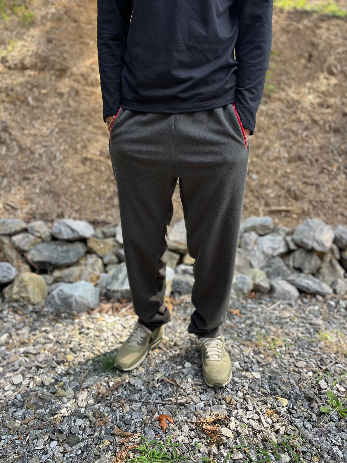 A person standing outdoors on gravel in front of a stone wall, wearing dark charcoal gray jogger pants with red side stripes, a black long-sleeve top, and olive green sneakers.