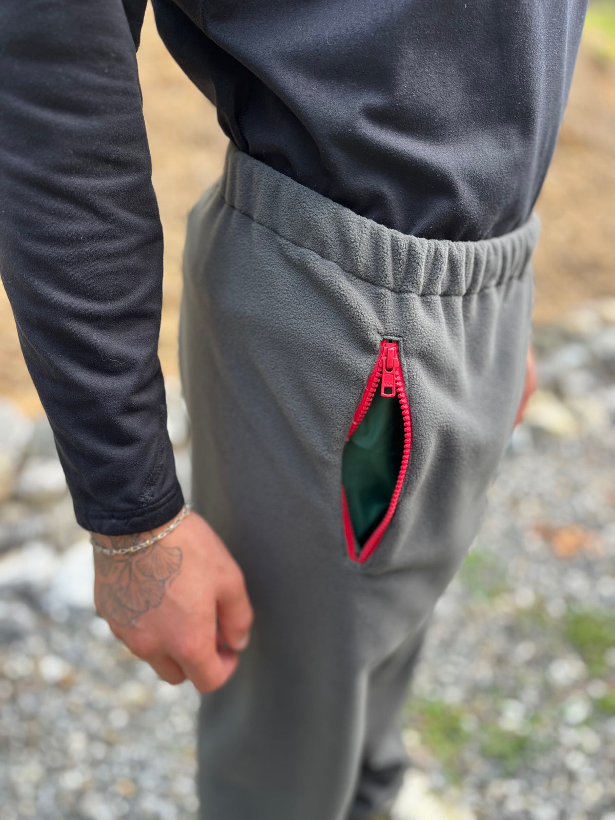 Close-up of a person wearing gray fleece jogger-style pants with a red zipper pocket, shown outdoors on a gravel surface.