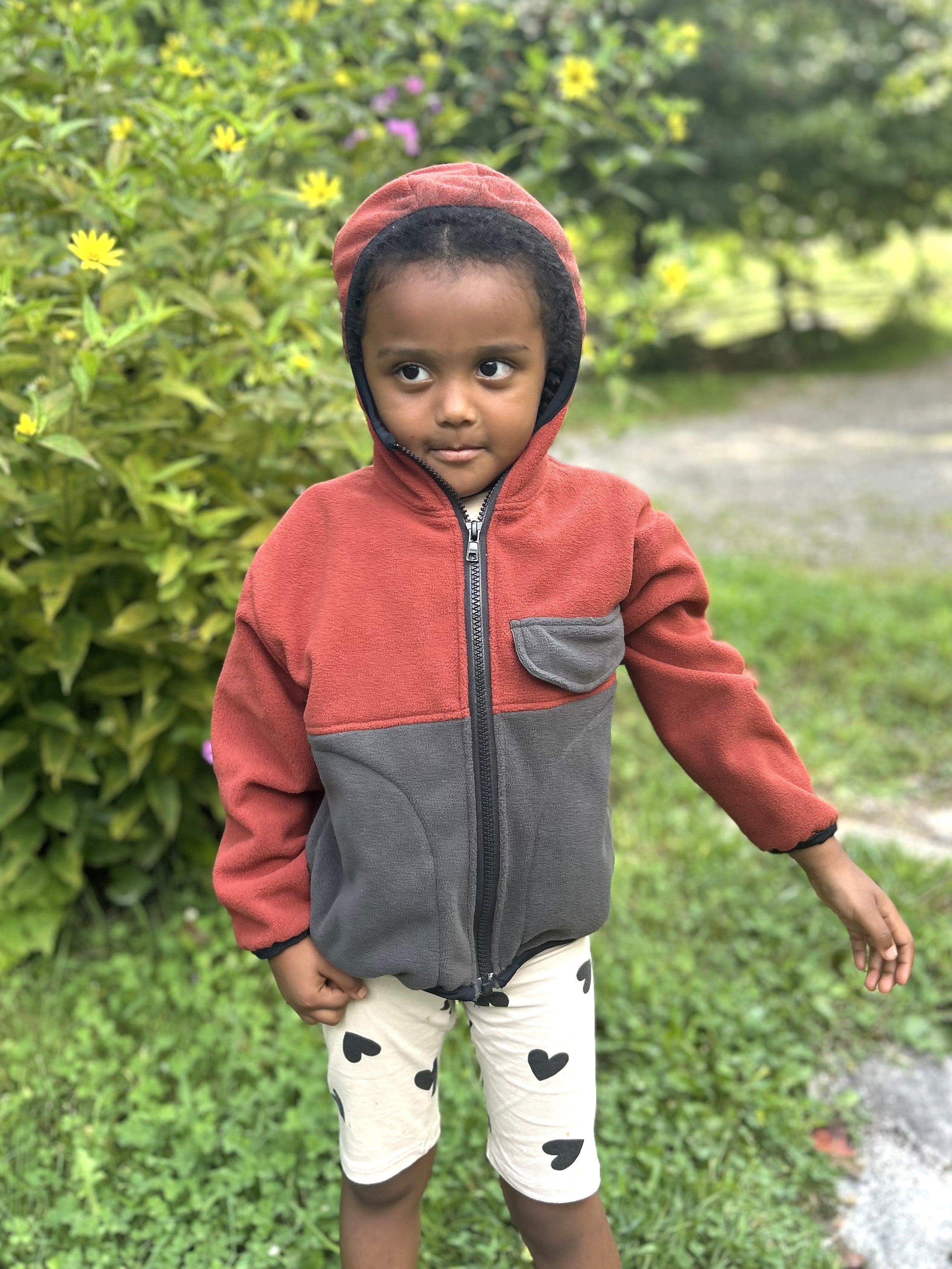 Young child with black braids in a red and grey zip up fleece hoodie standing outside
