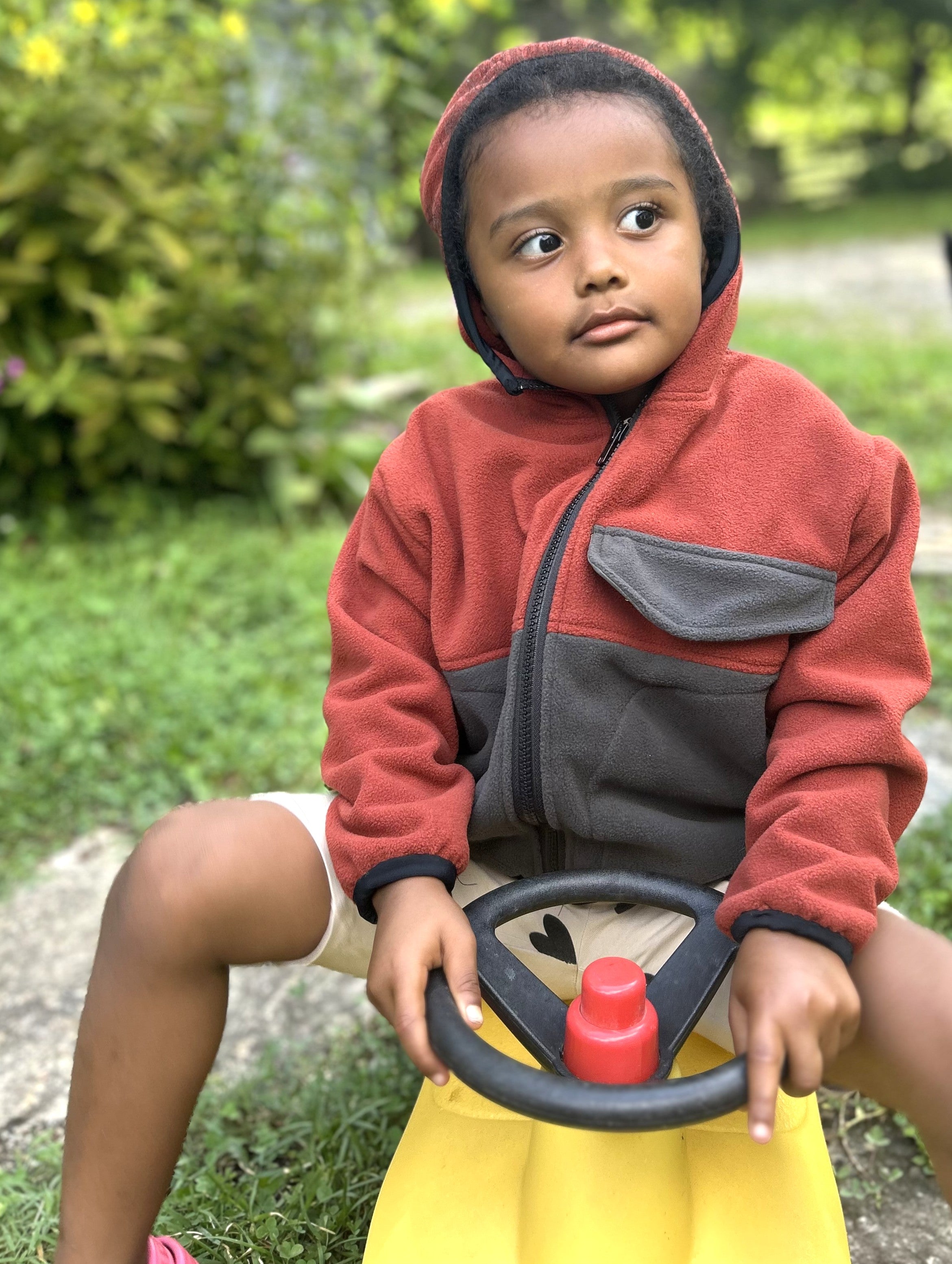Young child with black braids in a red and grey zip up fleece hoodie sitting on a yellow toy truck
