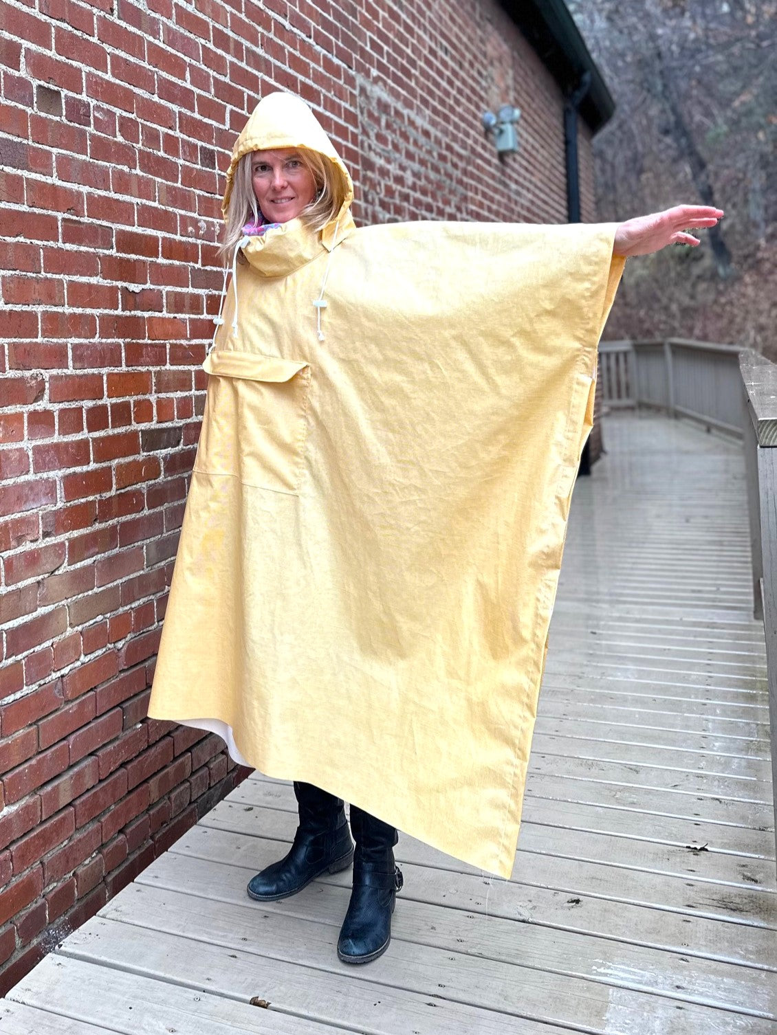 Woman standing a yellow rain poncho by a brick wall with her arm to the side to show how wide the poncho is.