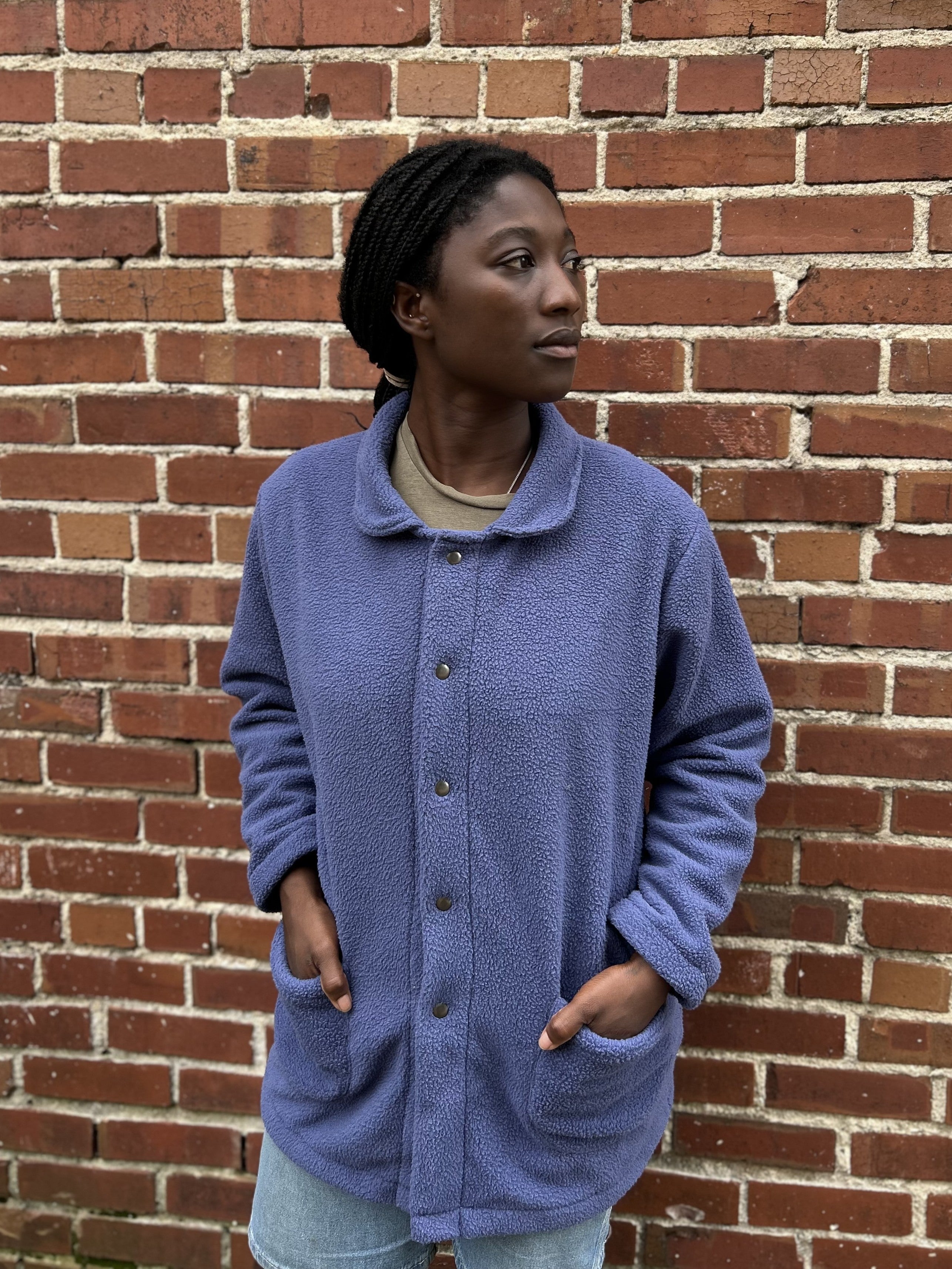 Woman standing in a purple fleece coat in front of a brick wall, with hands in pockets.