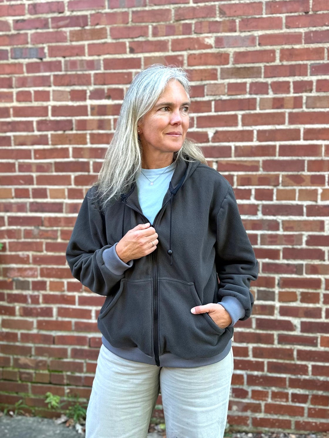 woman wearing a grey fleece zip up sweatshirt outside by a brick wall