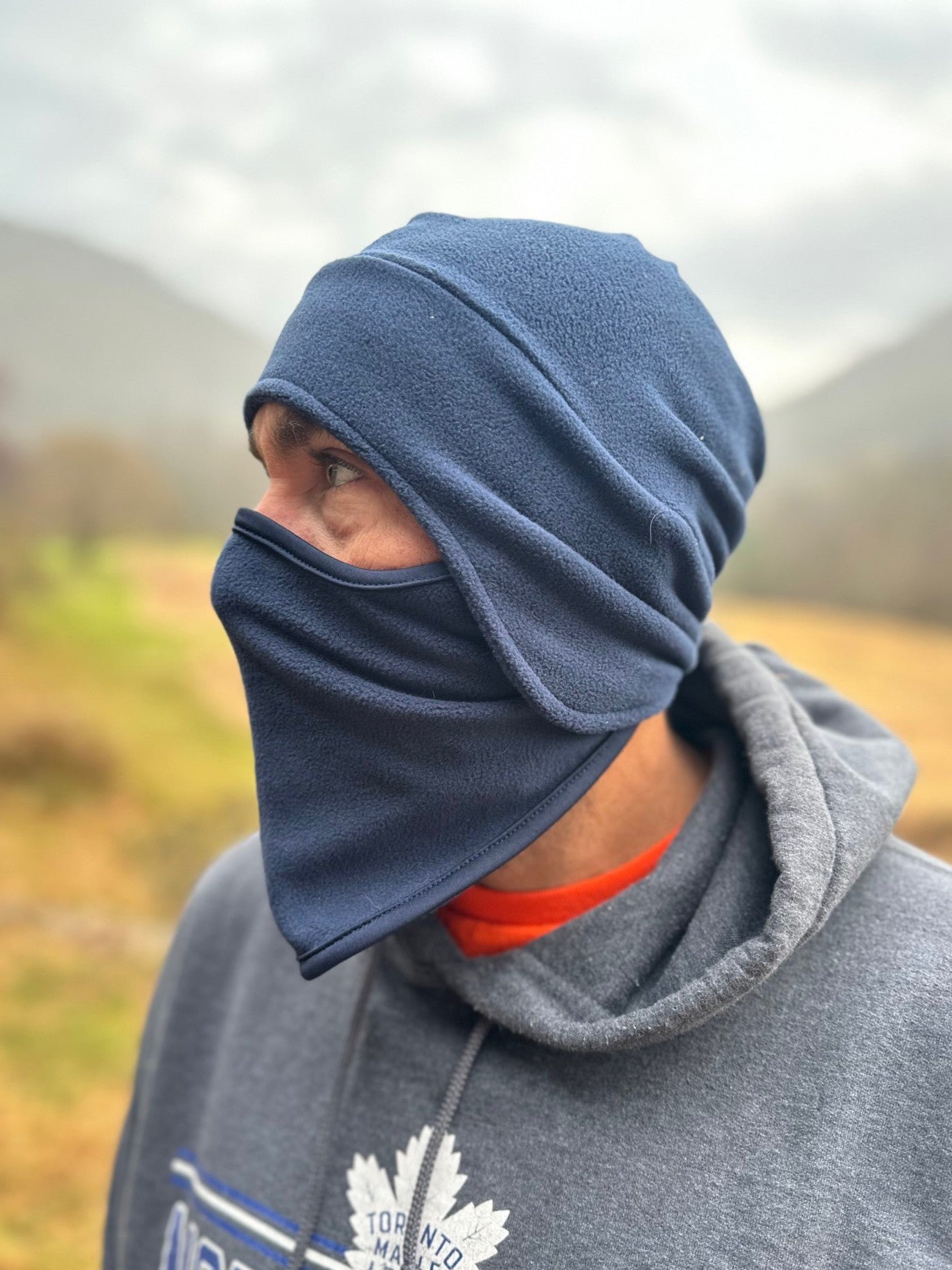 Person wearing a blue balaclava and gray hoodie with a logo in an outdoor setting