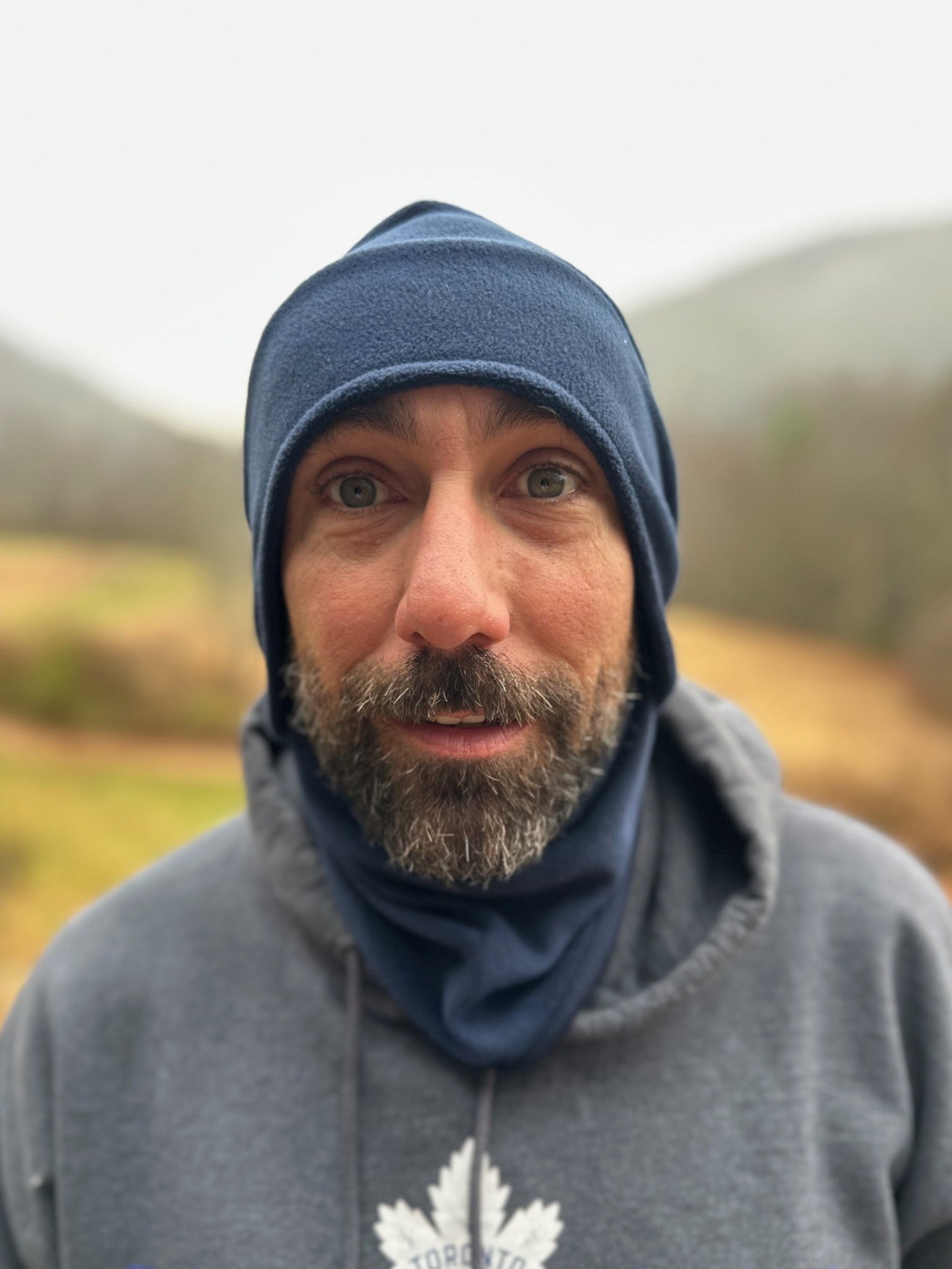 Man wearing a blue beanie and gray hoodie with a logo, outdoors.
