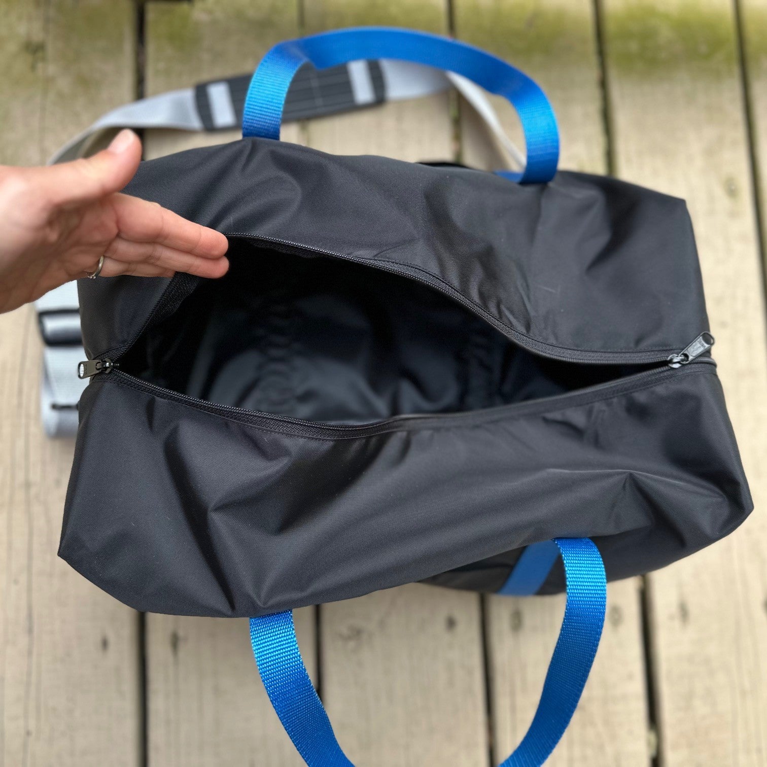 Black duffel bag with blue straps held open by a hand on a wooden surface