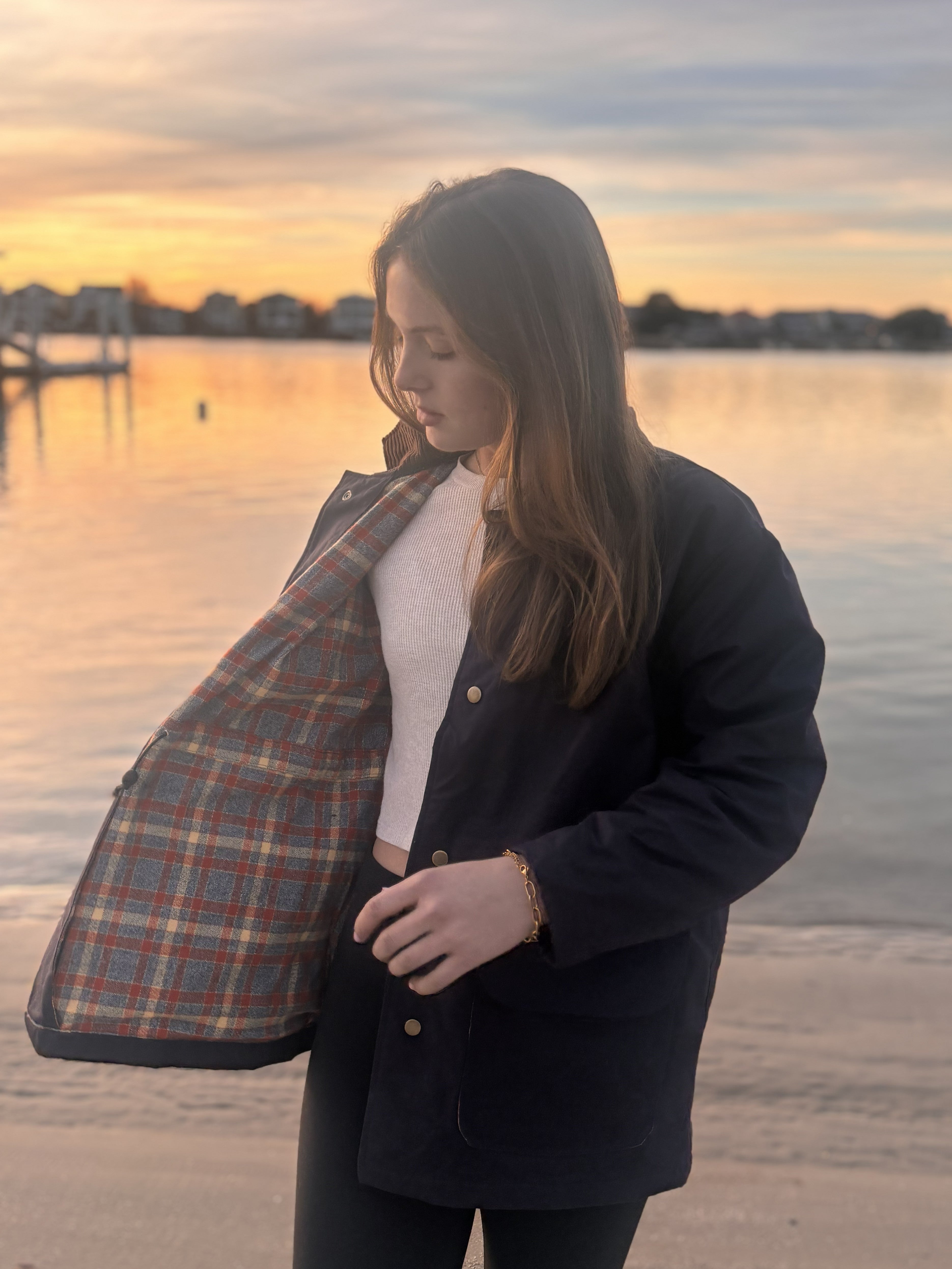 Woman holding a plaid jacket by a waterfront at sunset