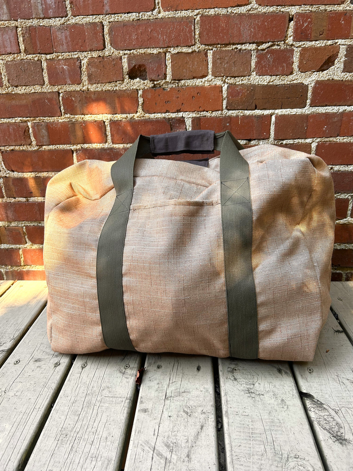 soft fabric suitcase in brown with olive webbing handles on a wooden deck and against a brick wall.
