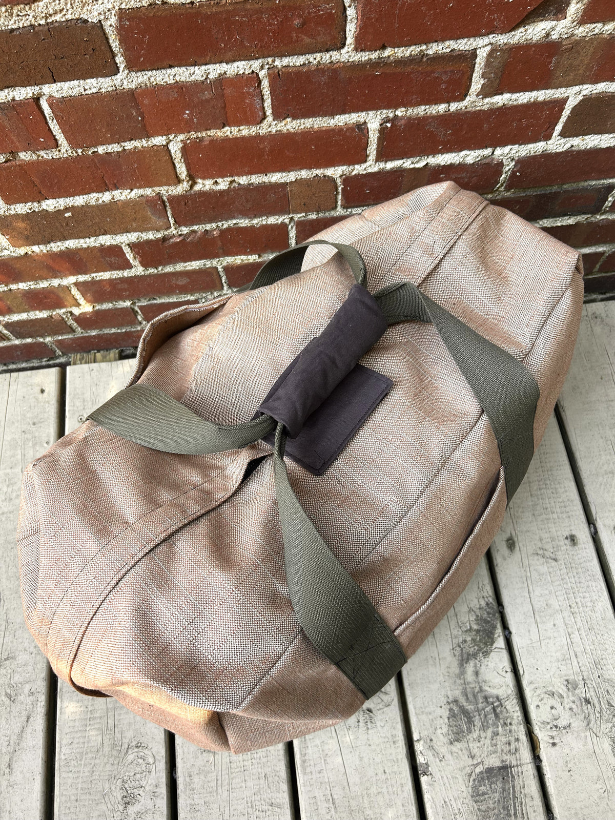 soft fabric suitcase in brown with olive webbing handles on a wooden deck and against a brick wall. View from top of suitcase