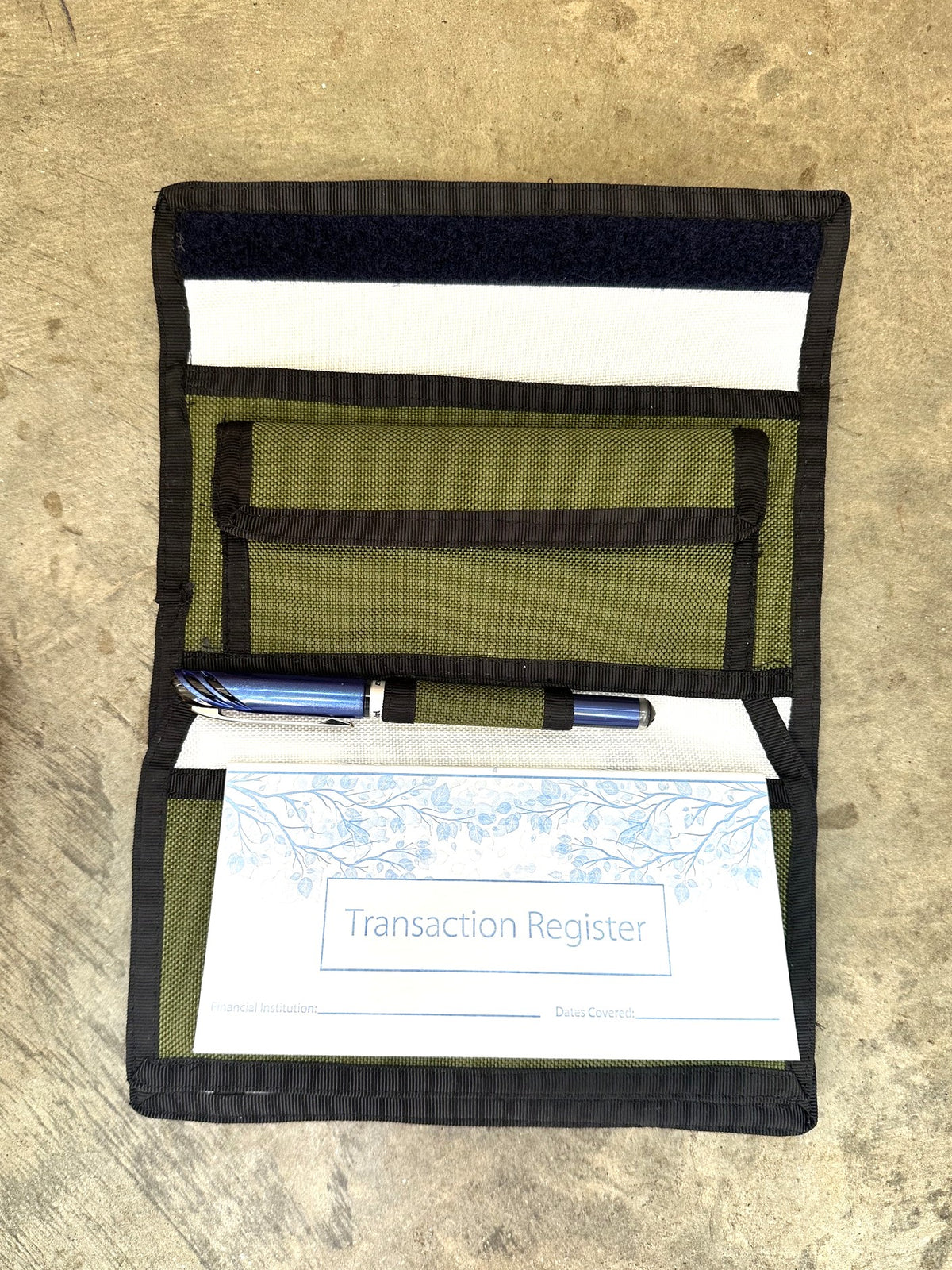 green checkbook wallet with black trim opened up with a pen on a concrete floor