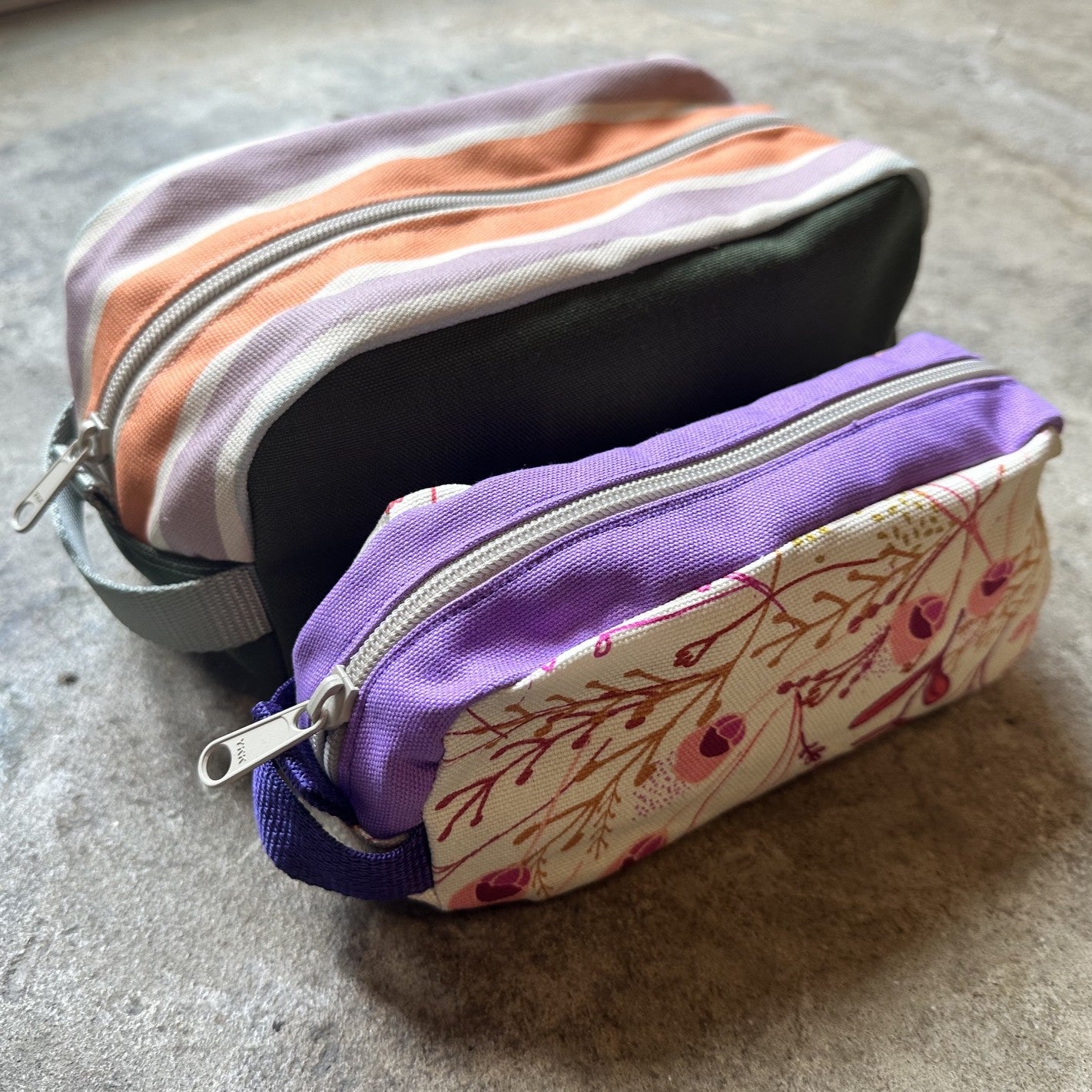 Two fabric zippered pouches resting on a textured gray surface. The larger pouch has horizontal stripes in lavender, peach, white, and dark green, with a gray strap.