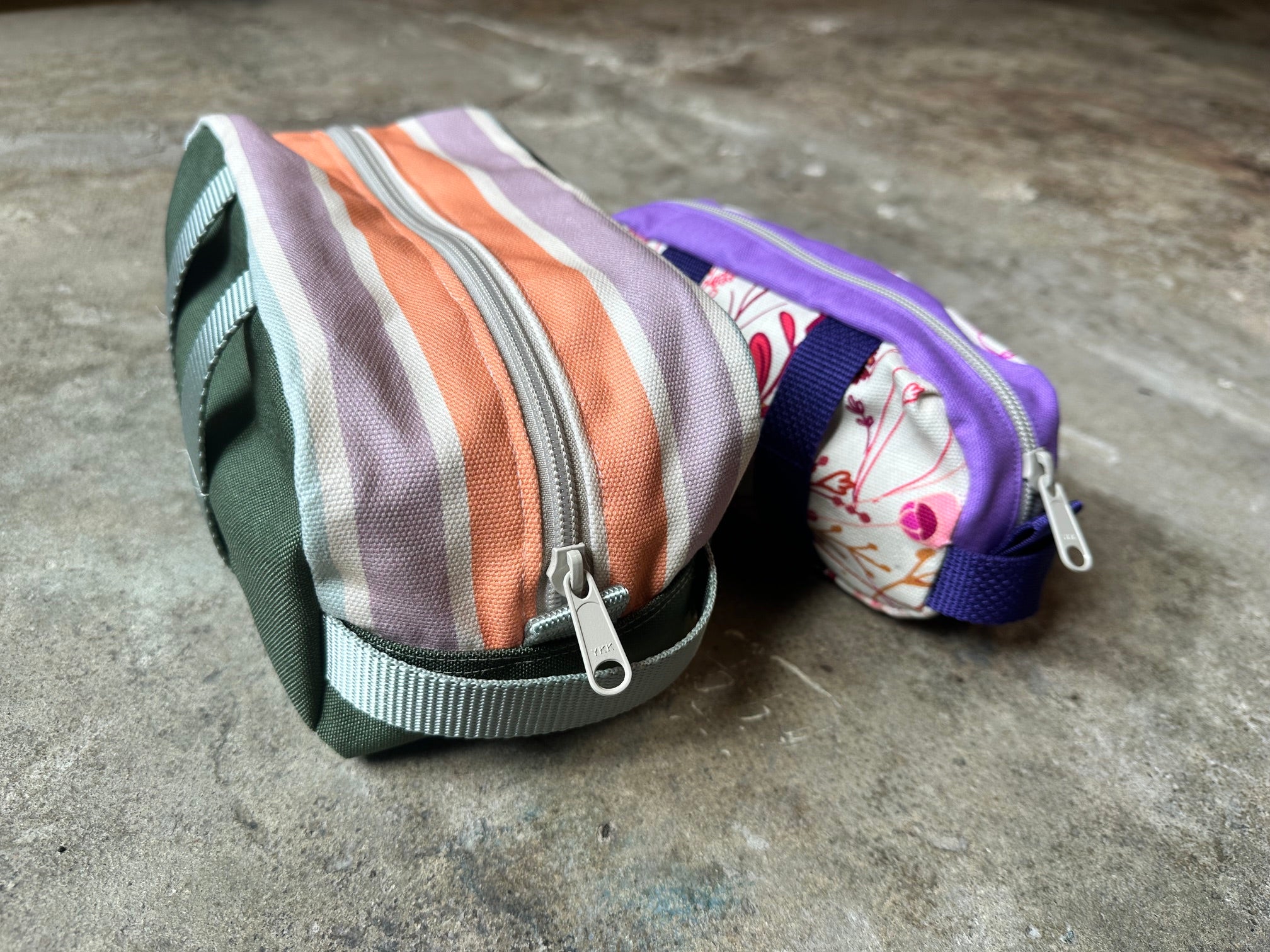 Two fabric zippered pouches resting on a textured concrete floor. The larger pouch in the foreground features vertical stripes in olive green, lavender, peach, and white, with a gray webbing strap.