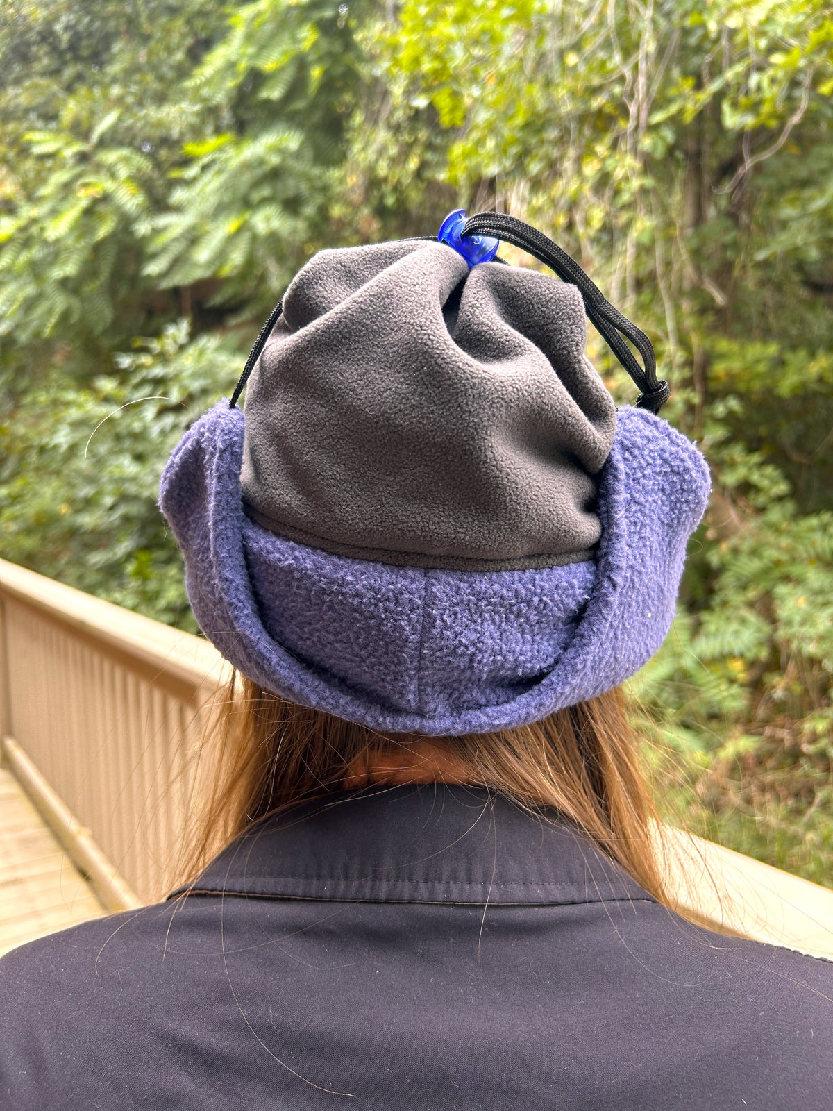 A person viewed from behind wearing a gray fleece winter hat with purple ear flaps and a black drawstring, standing outdoors on a wooden walkway with green foliage in the background.