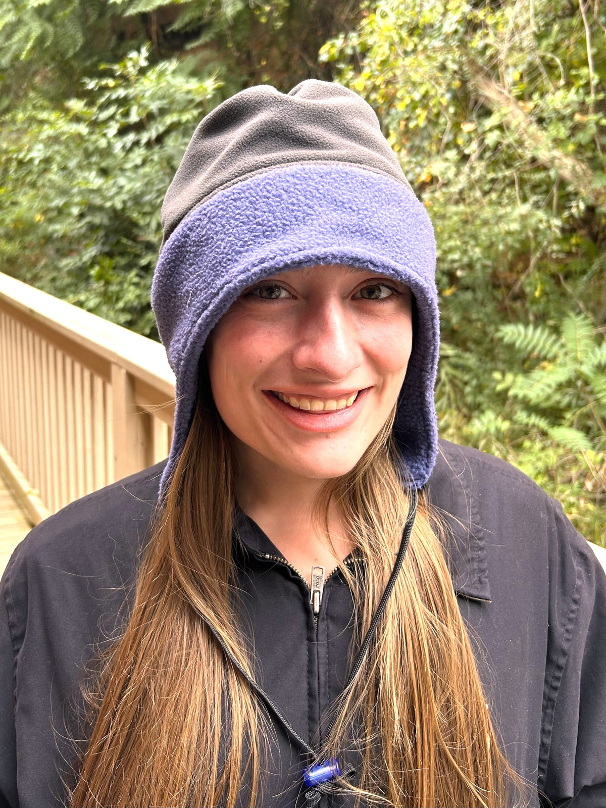 Woman standing outside by a fence and trees wearing a dark jacket and a two-color (blue and grey) toboggan.