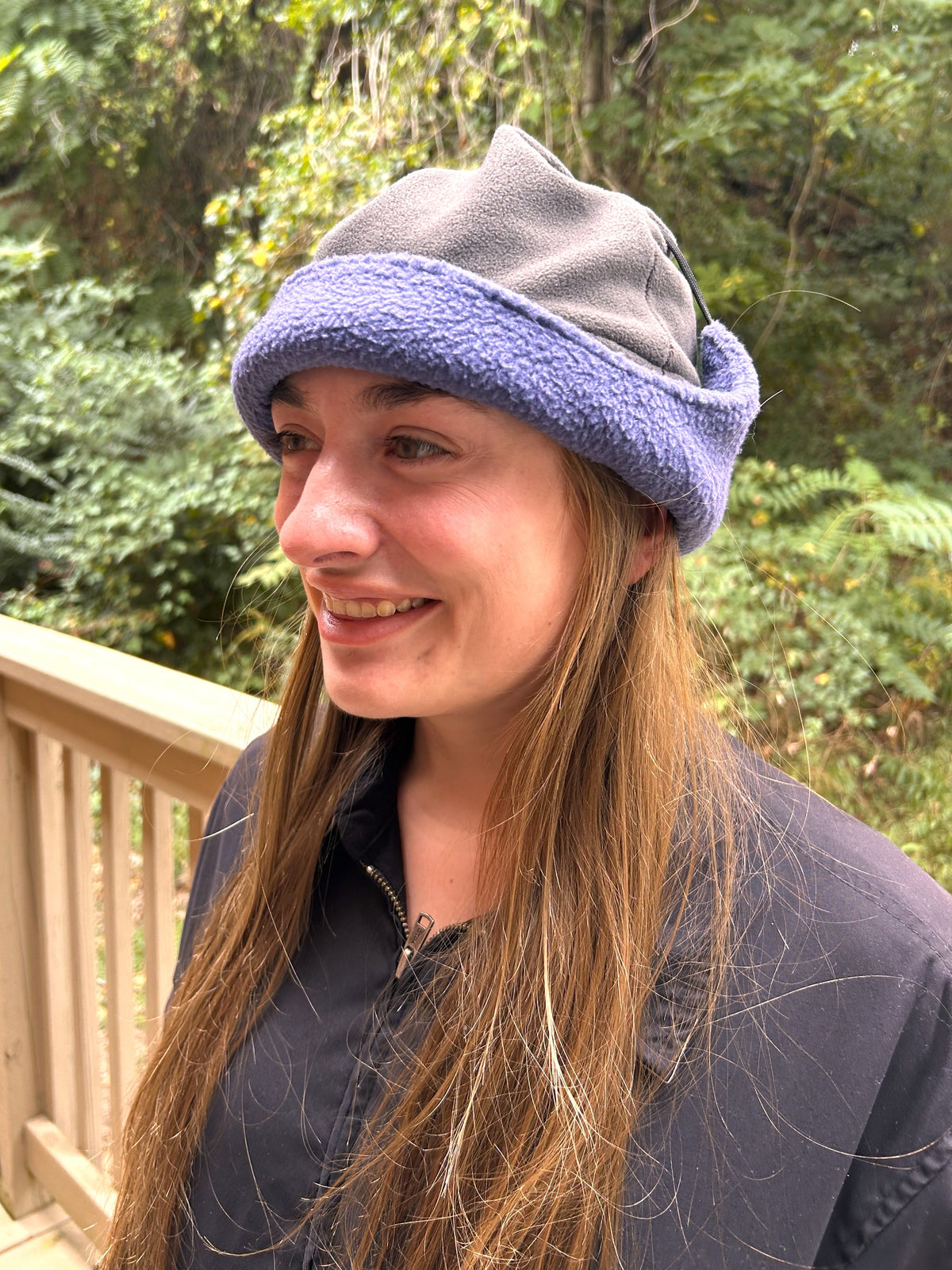 A woman wearing a two-tone fleece beanie hat with a gray top and a lavender-purple folded brim, photographed outdoors on a wooden deck with green foliage in the background.