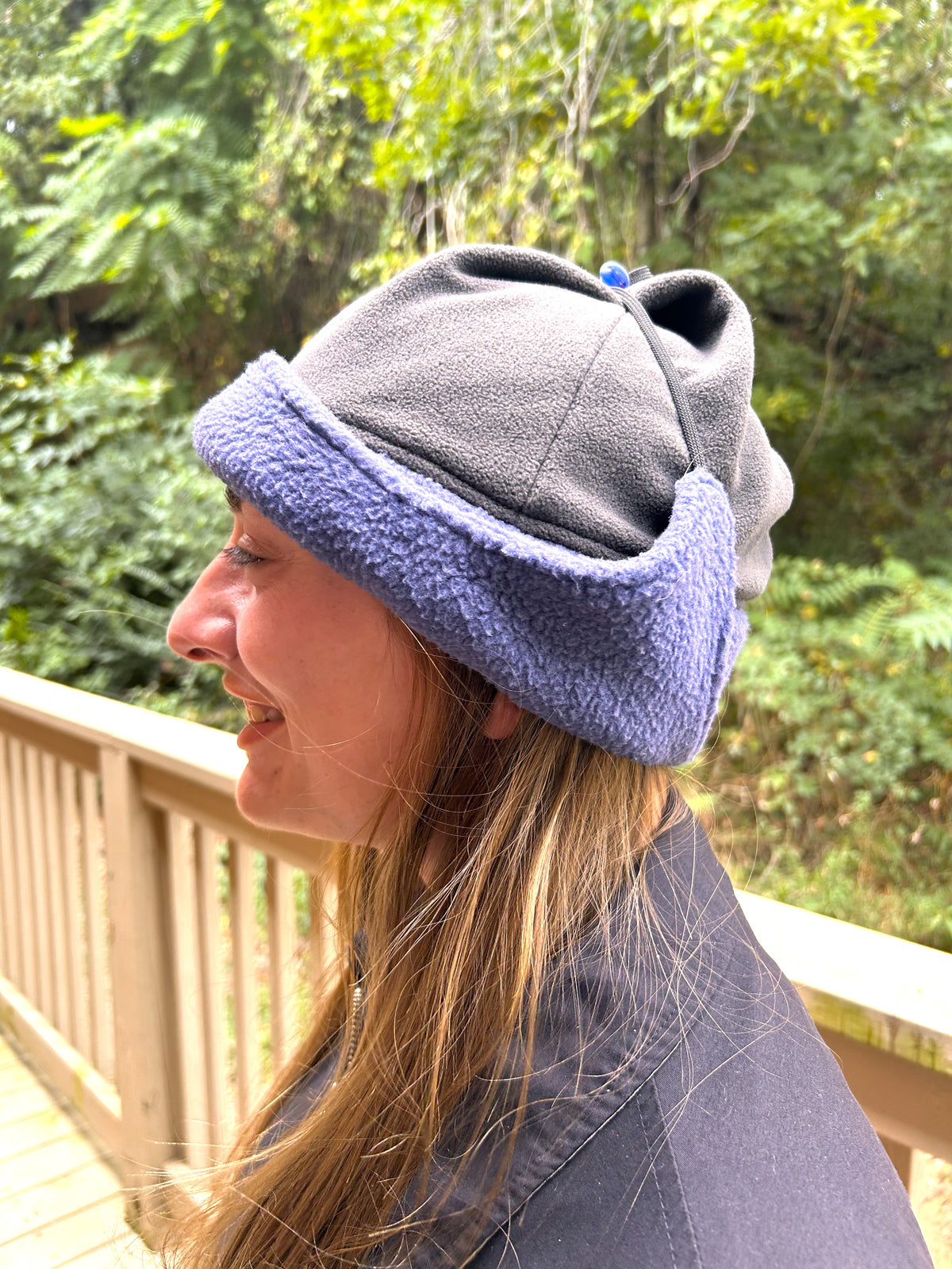 A woman in profile wearing a gray fleece beanie with a light purple fleece trim, standing on a wooden deck with a forest background.