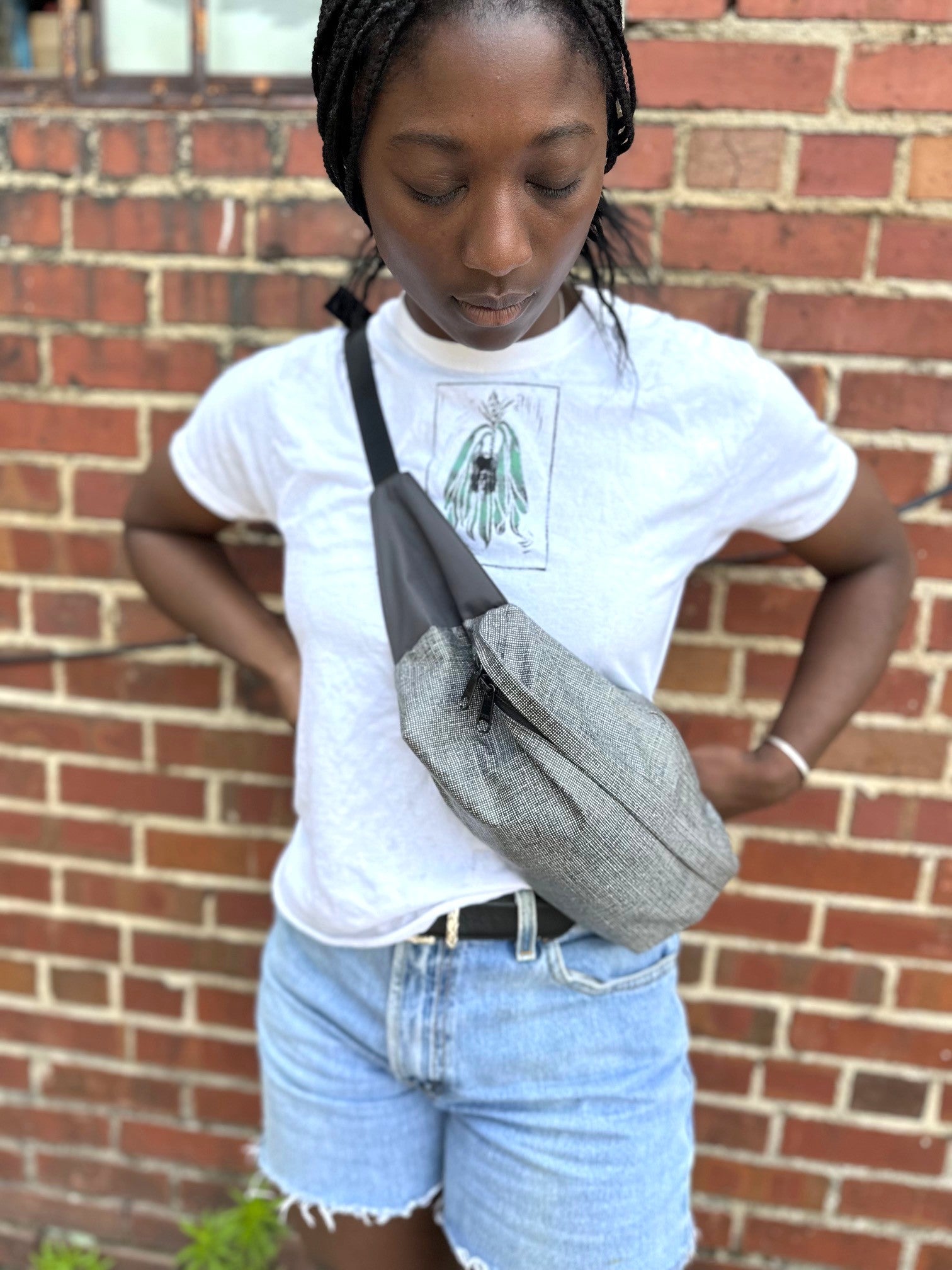 Person wearing a white t-shirt with a graphic design and denim shorts, holding a gray crossbody bag against a brick wall.