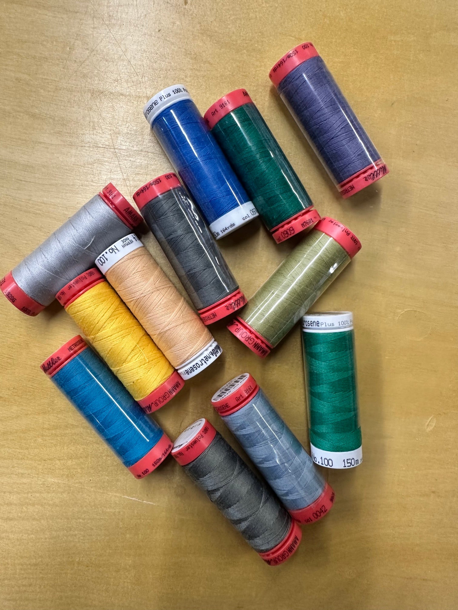 A group of 12 spools of sewing thread in various colors, including blue, green, gray, yellow, beige, and purple, arranged on a light wood surface.