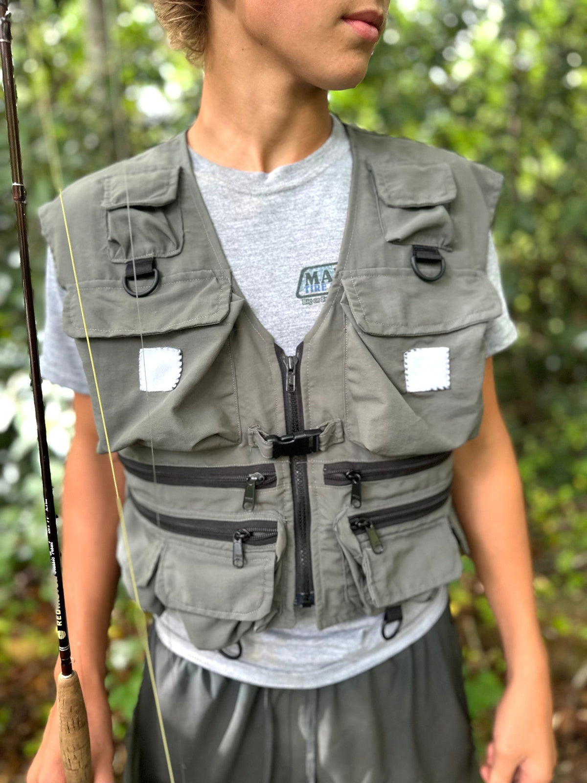 Close up of young man wearing grey shorts, shirt, and fishing vest in the woods and carrying a fly rod