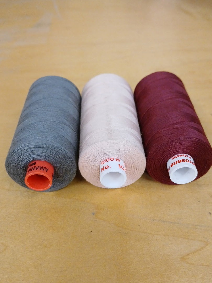 Three spools of sewing thread in gray, light pink, and burgundy, arranged side by side on a light wood surface.