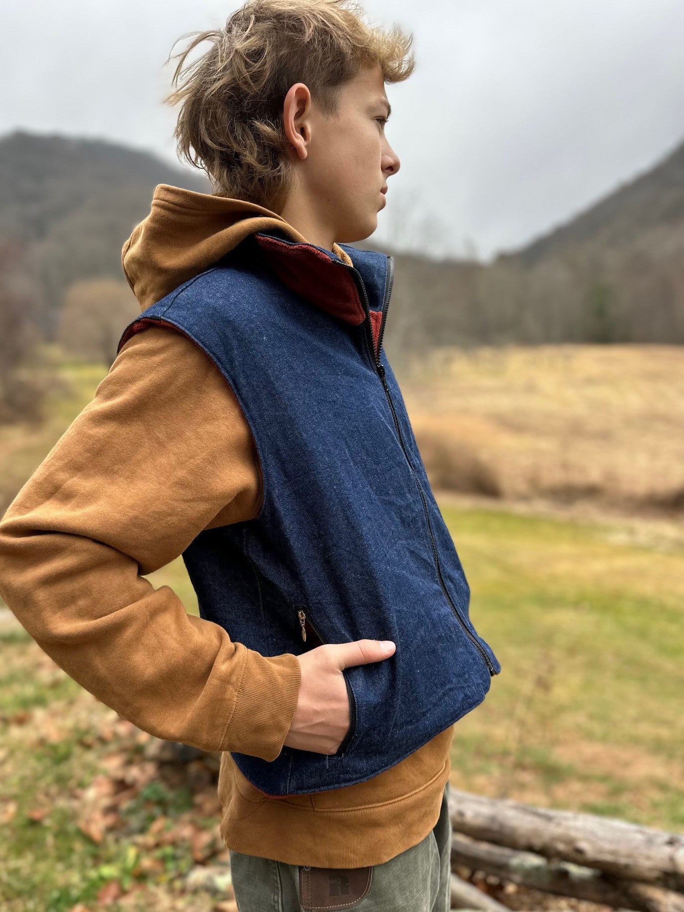 Person wearing a brown sweatshirt and blue vest standing outdoors with mountains in the background