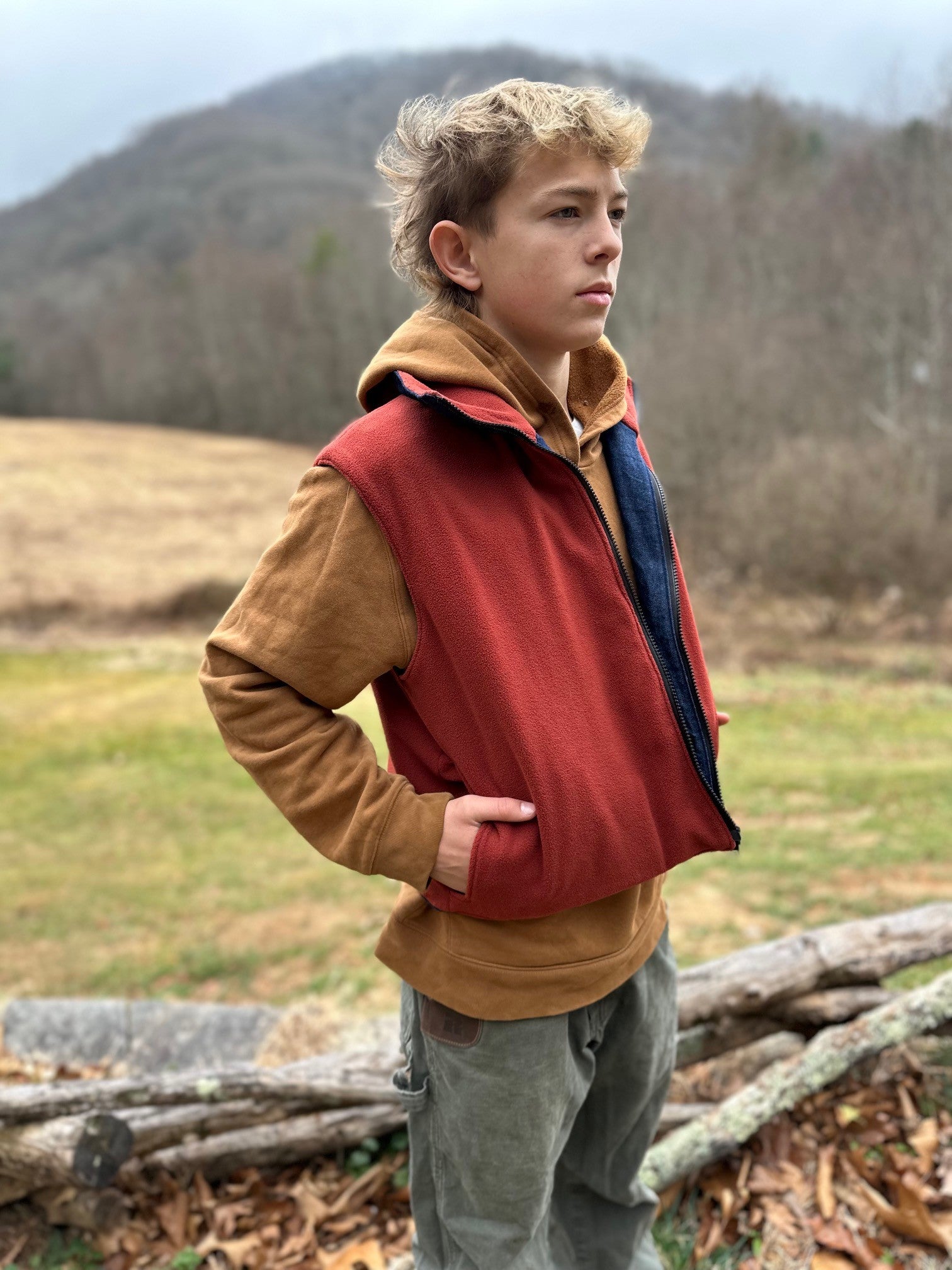 Person wearing a red vest over a brown jacket in a natural setting with mountains in the background.