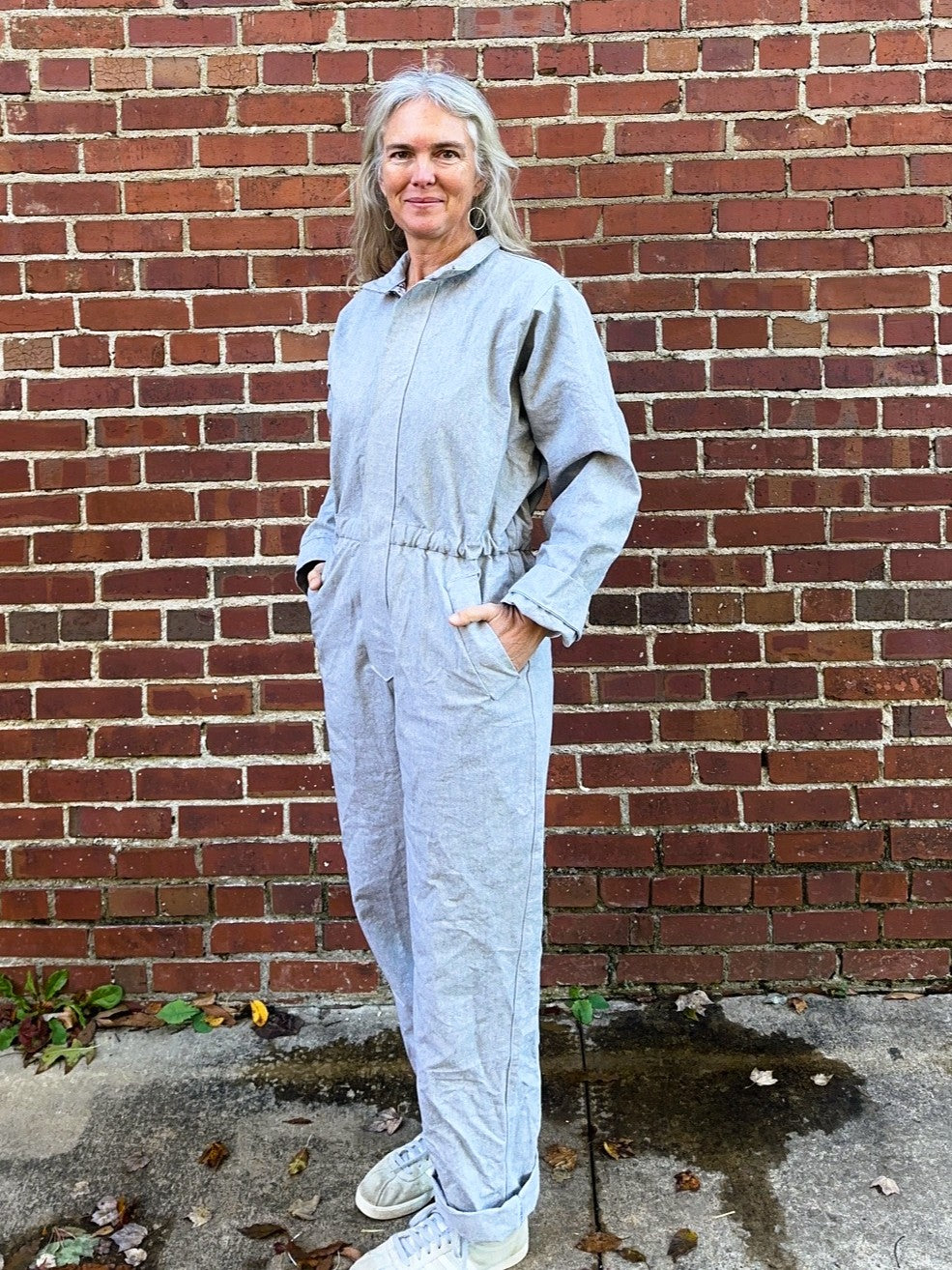 Person wearing a light gray jumpsuit standing against a brick wall.