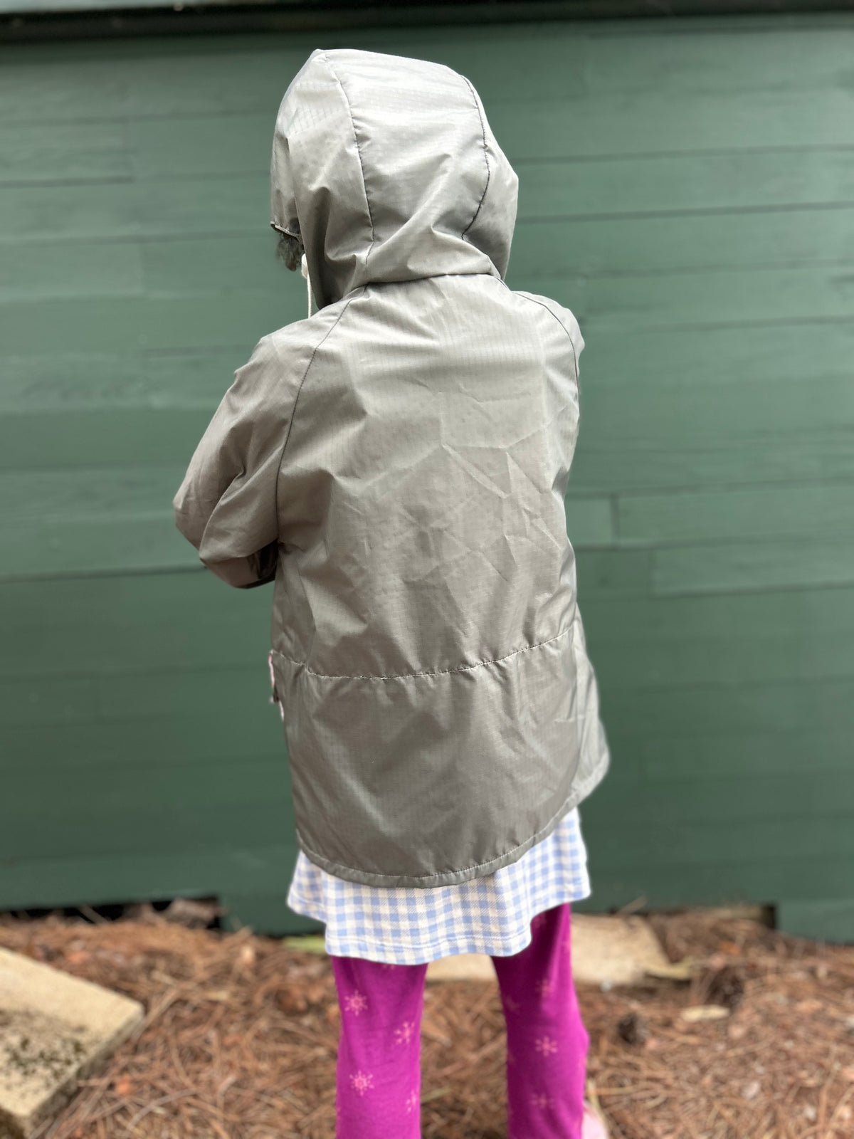 A child wearing a hooded light gray jacket, shown from the back, standing outdoors in front of a green wooden wall.