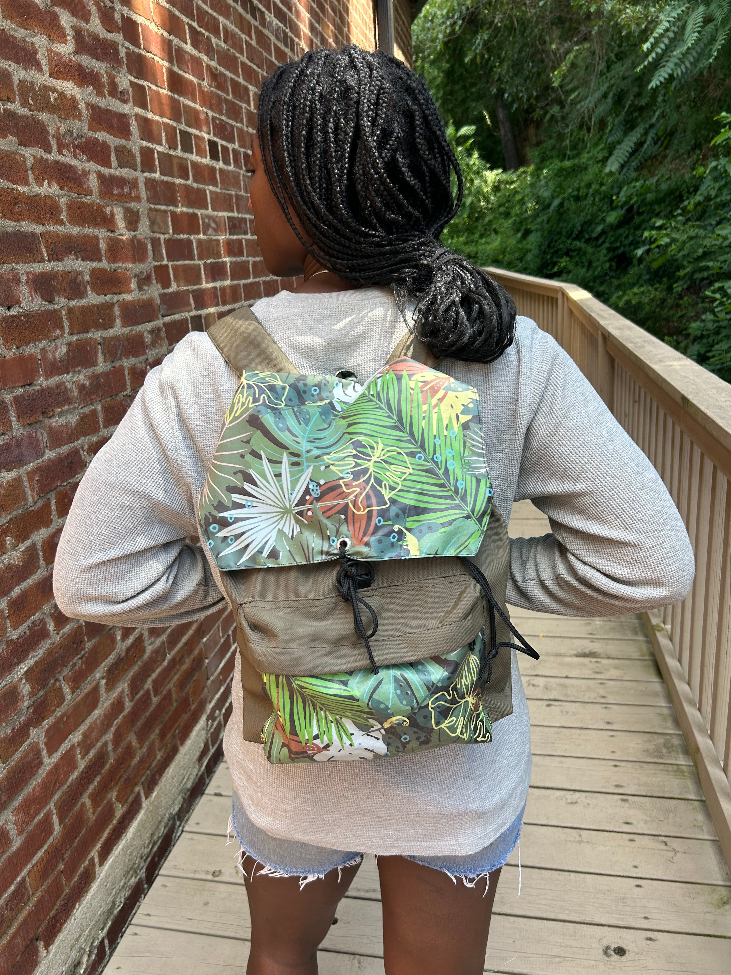 A person wearing a light gray sweatshirt and denim shorts, carrying a beige backpack with a green tropical leaf print front panel and black drawstring closure, standing on a brick patio next to a wooden deck.