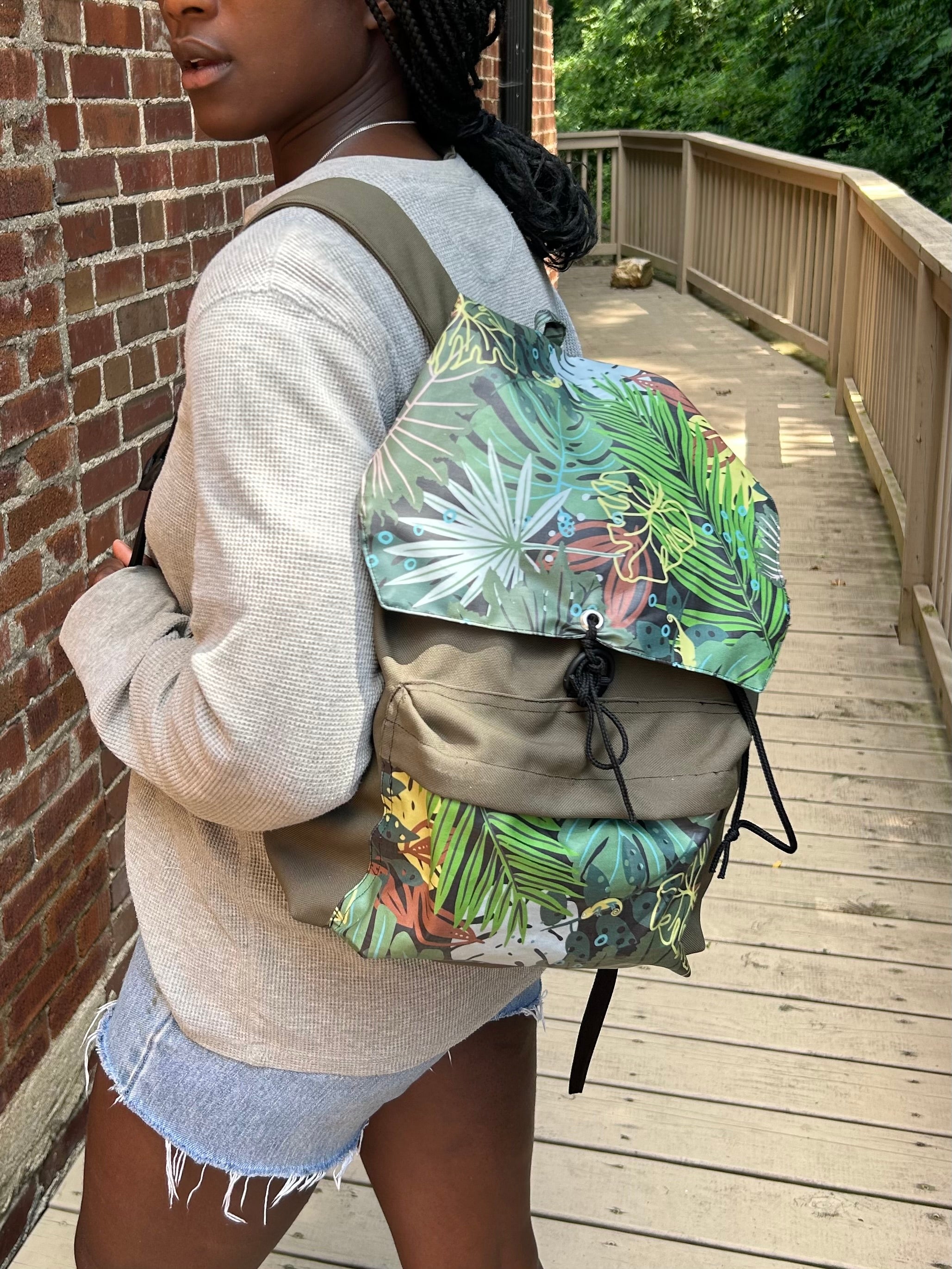 A woman wearing a light beige knit sweater and denim shorts carries a backpack with a green tropical palm leaf pattern and olive green fabric panels, walking on a wooden boardwalk next to a brick wall.