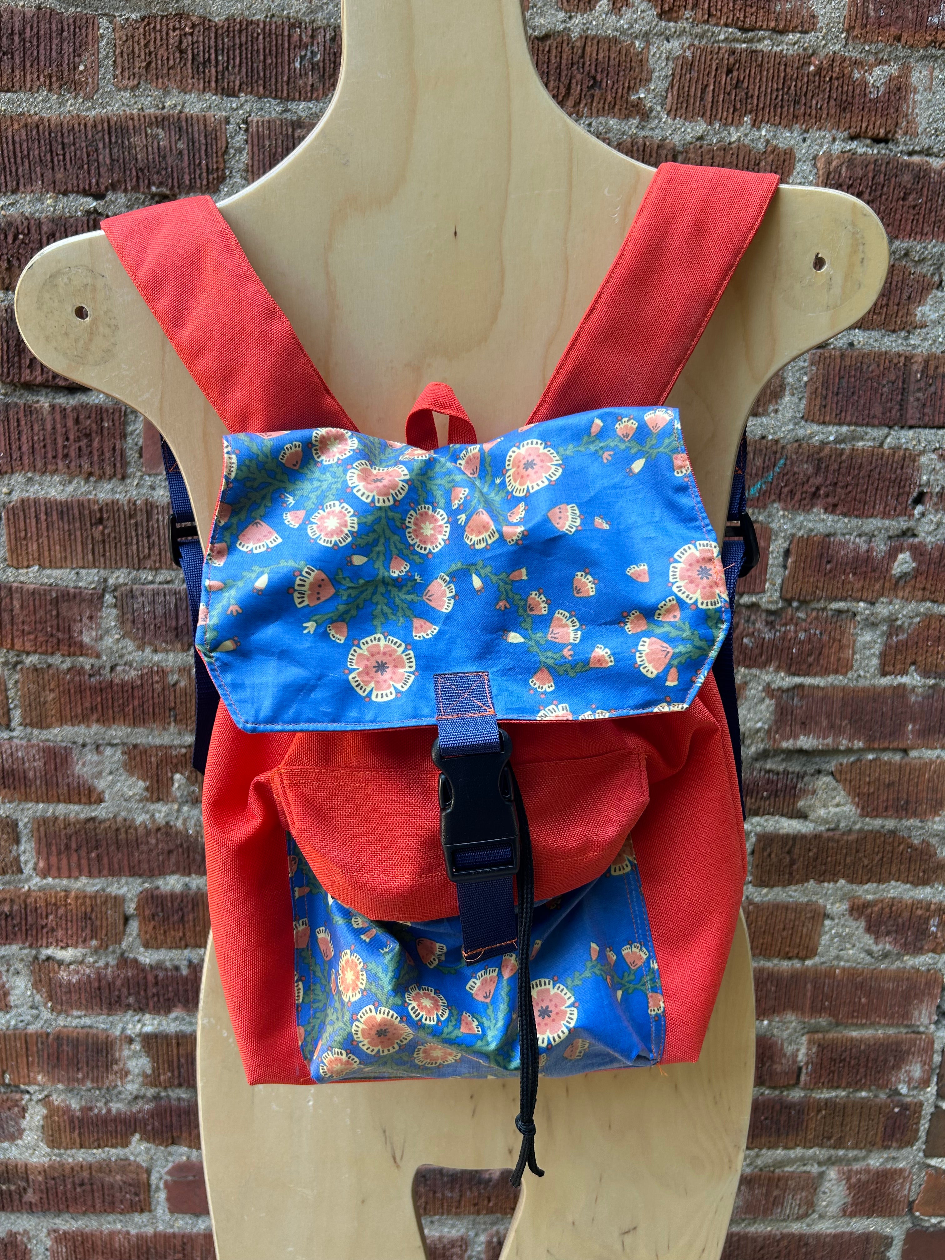 A small backpack with bright red fabric sides and straps, and blue floral front panels, displayed on a wooden mannequin torso against a brick wall background.