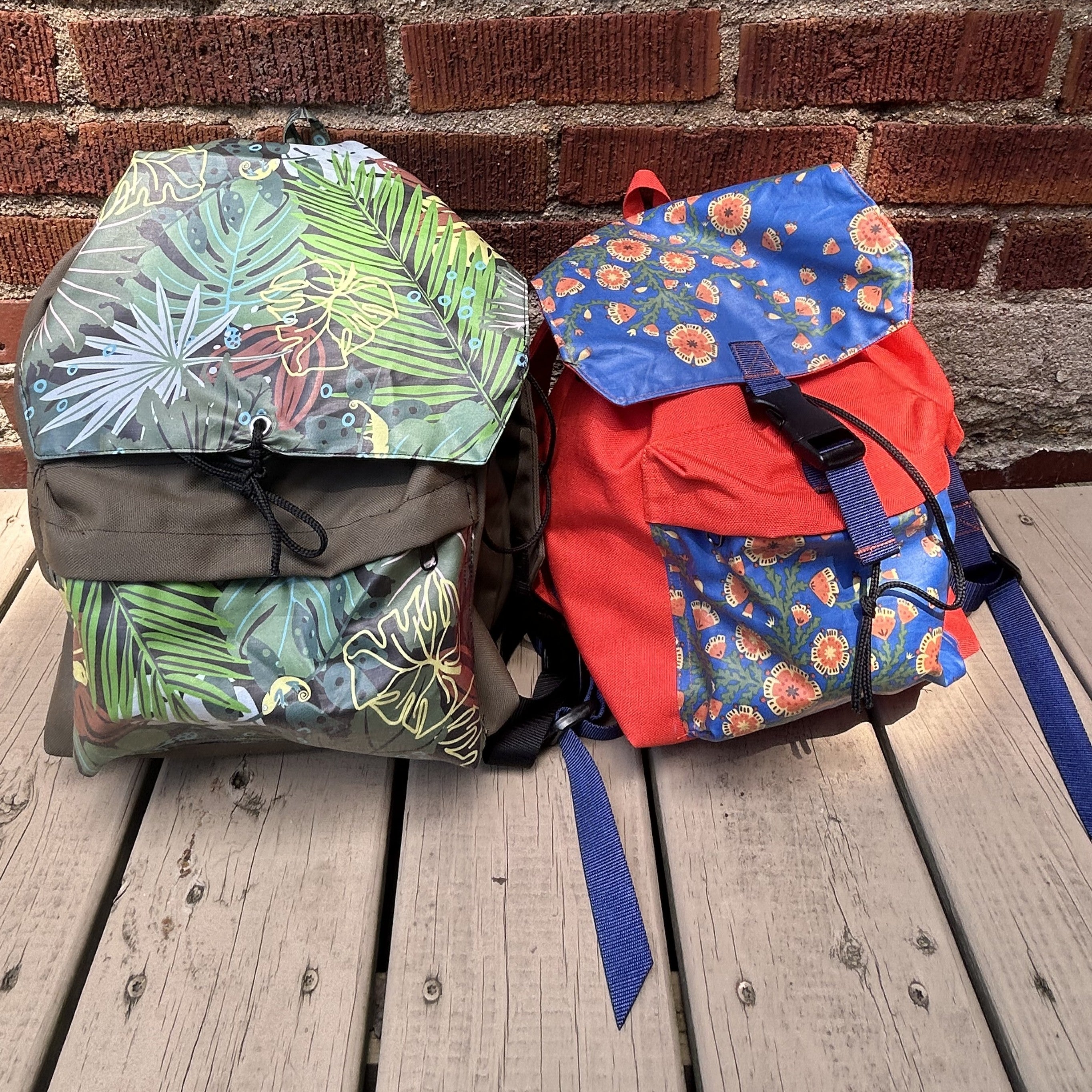 Two day packs with flip-top lids, one featuring a green tropical leaf print and the other a blue floral print with a bright red body, displayed on a wooden deck in front of a brick wall.