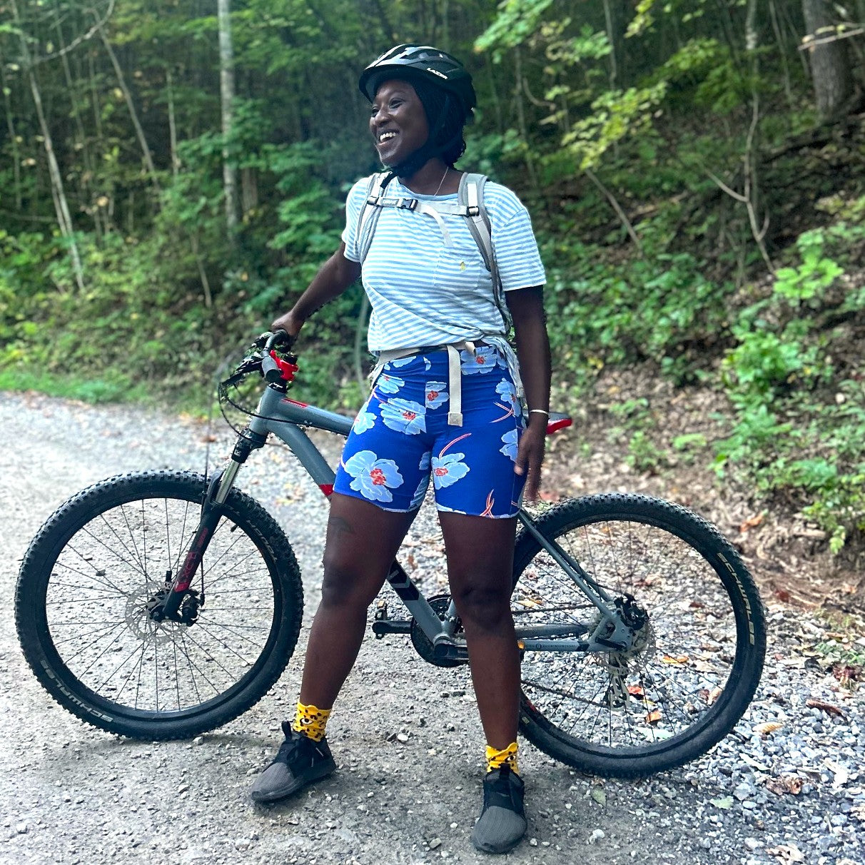 Person standing with a bicycle on a trail in a forest wearing blue floral biking shorts.