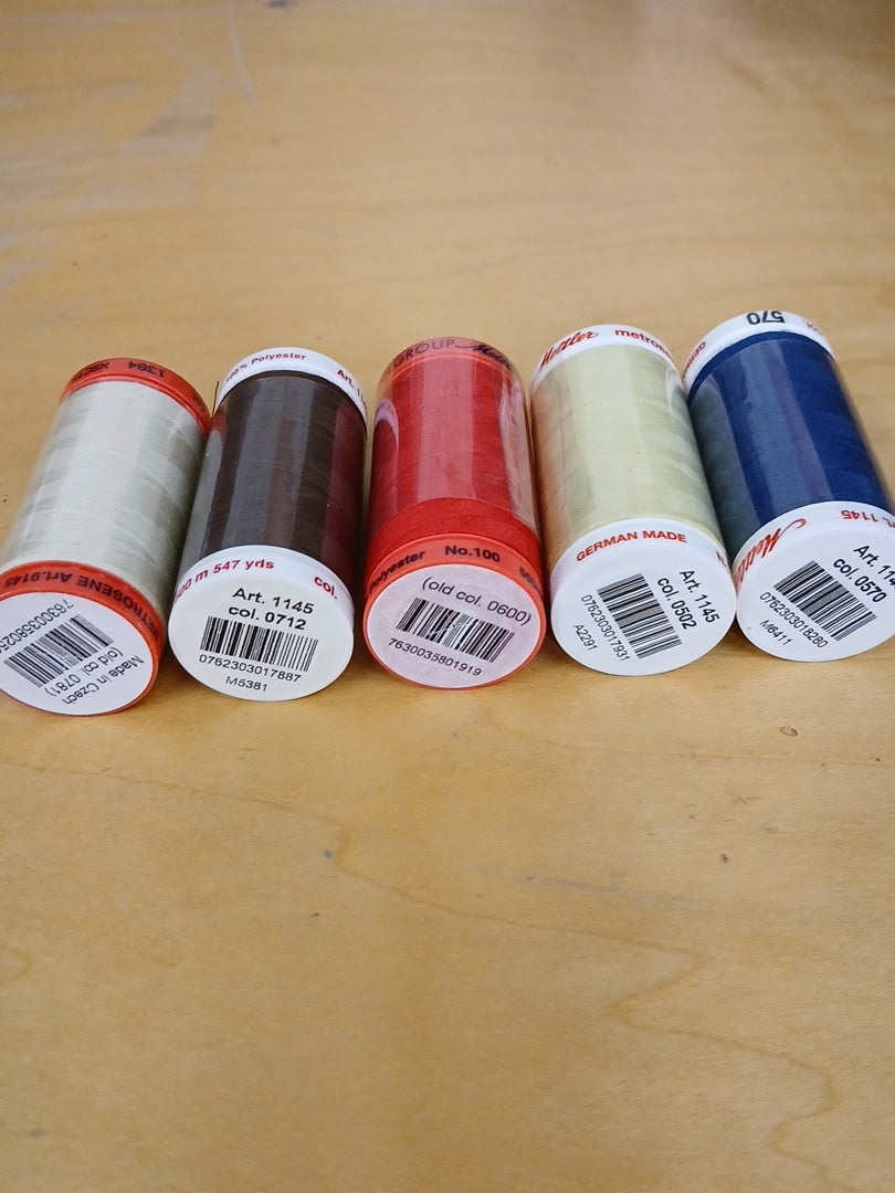 Five spools of thread in various colors lined up on a light wood surface. The spools include white, dark brown, red, cream, and blue threads, each with white labels showing barcodes and text.