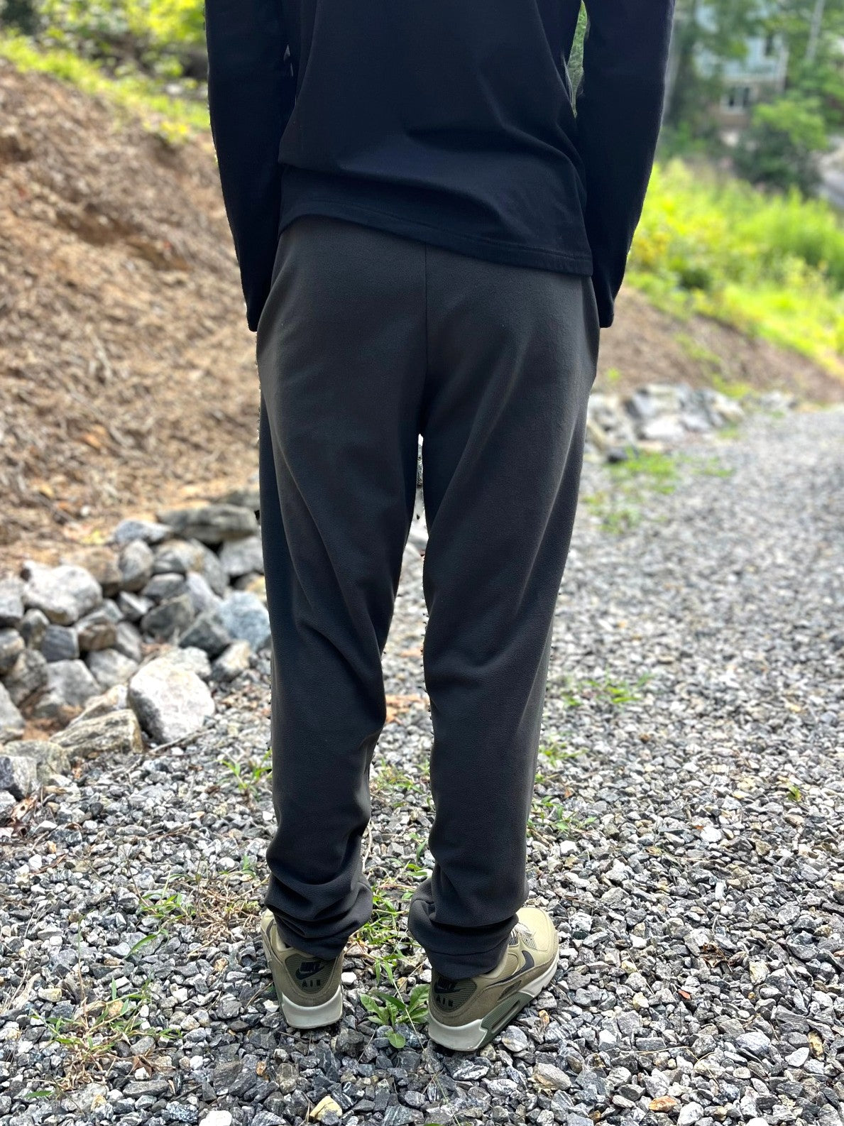 A person viewed from behind wearing dark charcoal gray jogger sweatpants, a black long-sleeve top, and olive green sneakers on a gravel path outdoors.