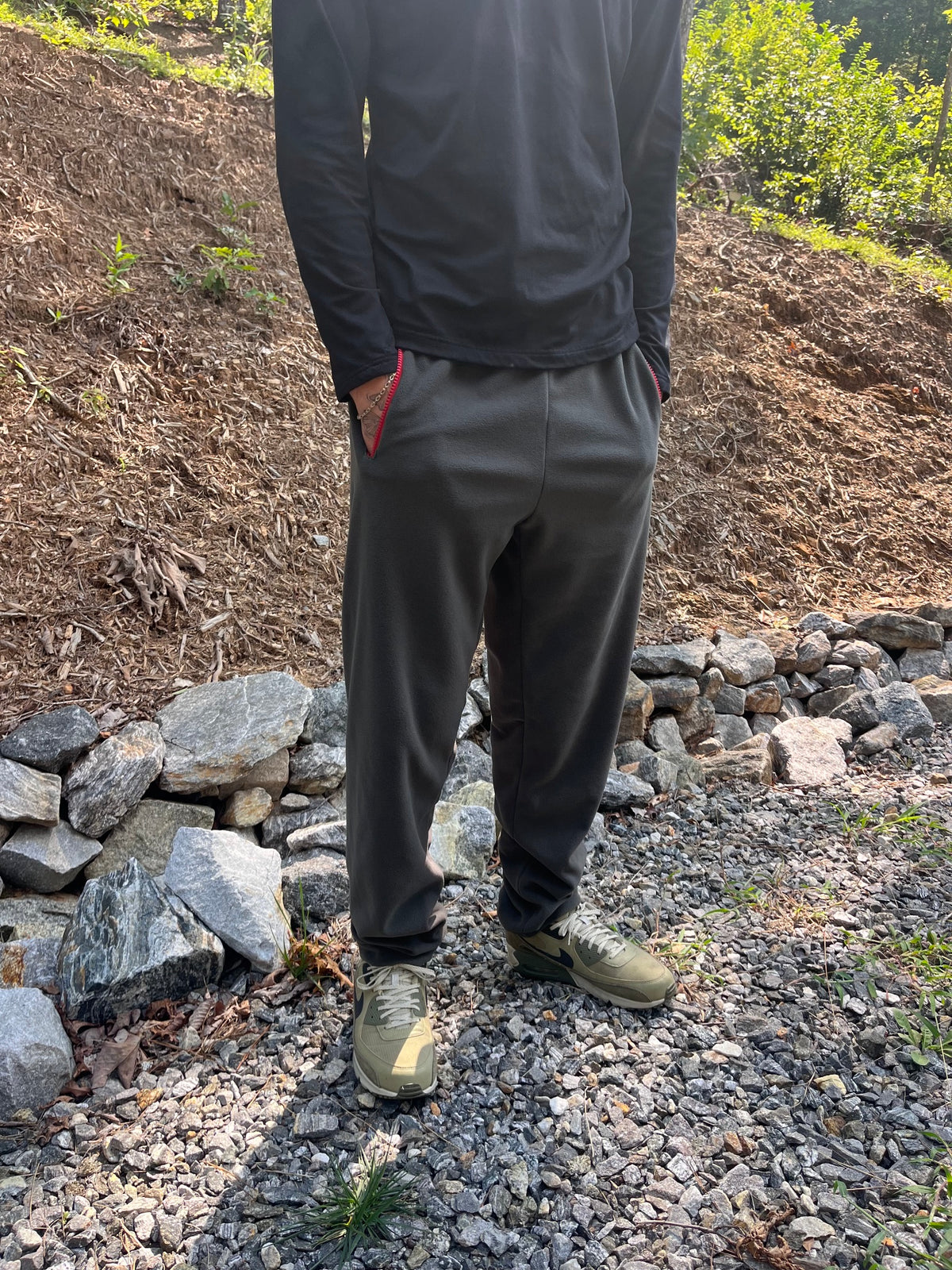A person standing outdoors on a gravel path wearing dark charcoal gray jogger pants, a black long-sleeve top, and olive green athletic shoes, with a dirt slope and rocks behind them.