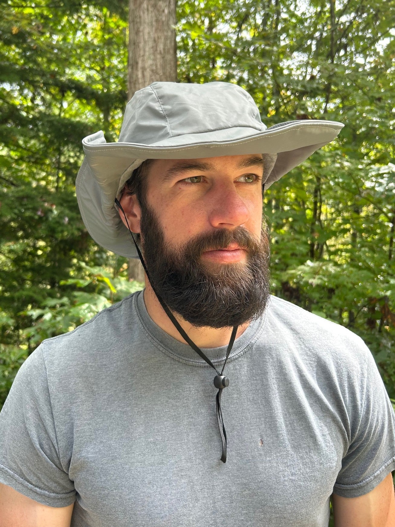 Man wearing a wide-brimmed grey hat  and grey tshirt outdoors with greenery in the background
