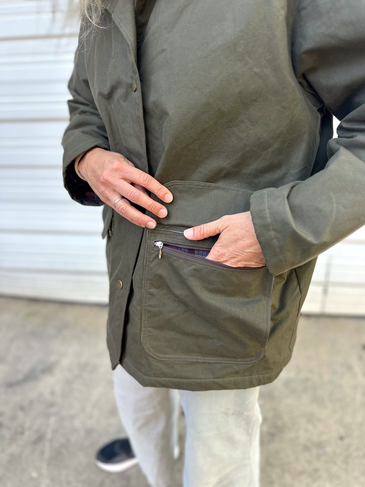 Close up of woman standing in front of a white garage door wearing tan pants and an olive barn jacket. Hand in pocket