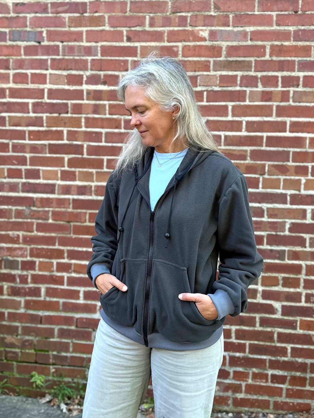 woman wearing a grey fleece zip up sweatshirt outside by a brick wall