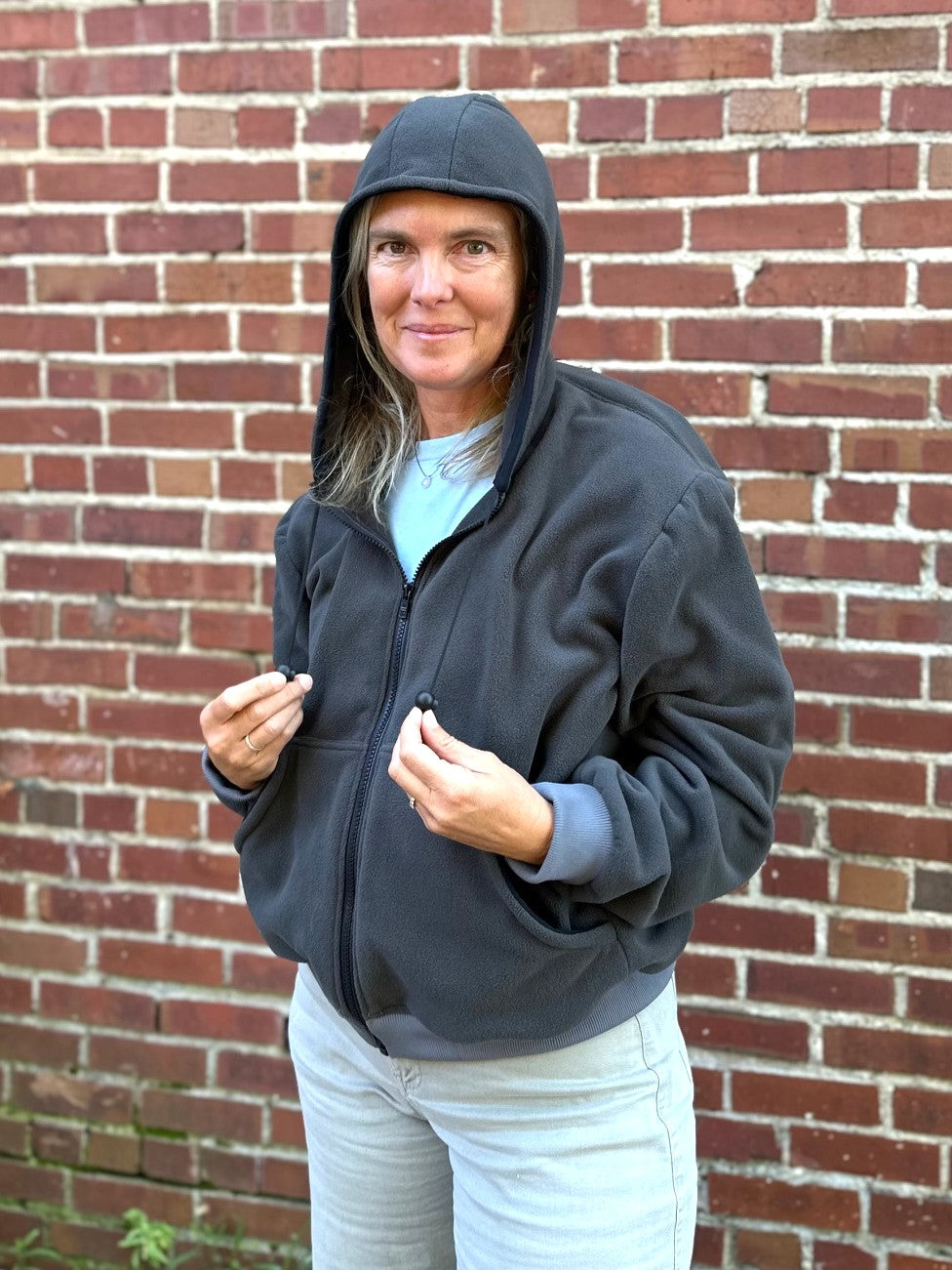 woman wearing a grey fleece zip up sweatshirt outside by a brick wall with hood up