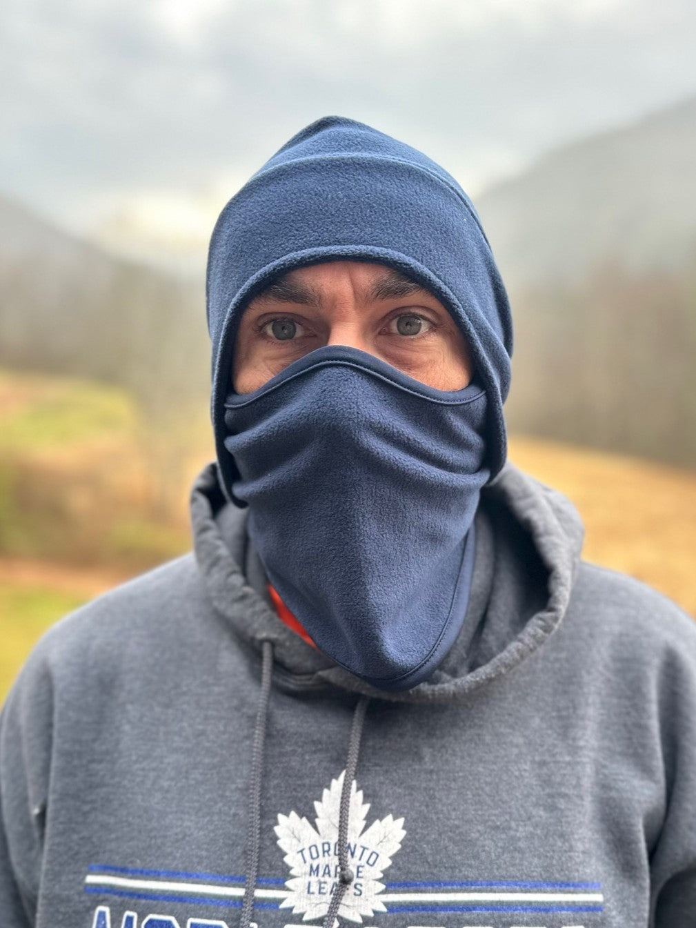 Person wearing a blue balaclava and gray hoodie with a logo, standing outdoors.