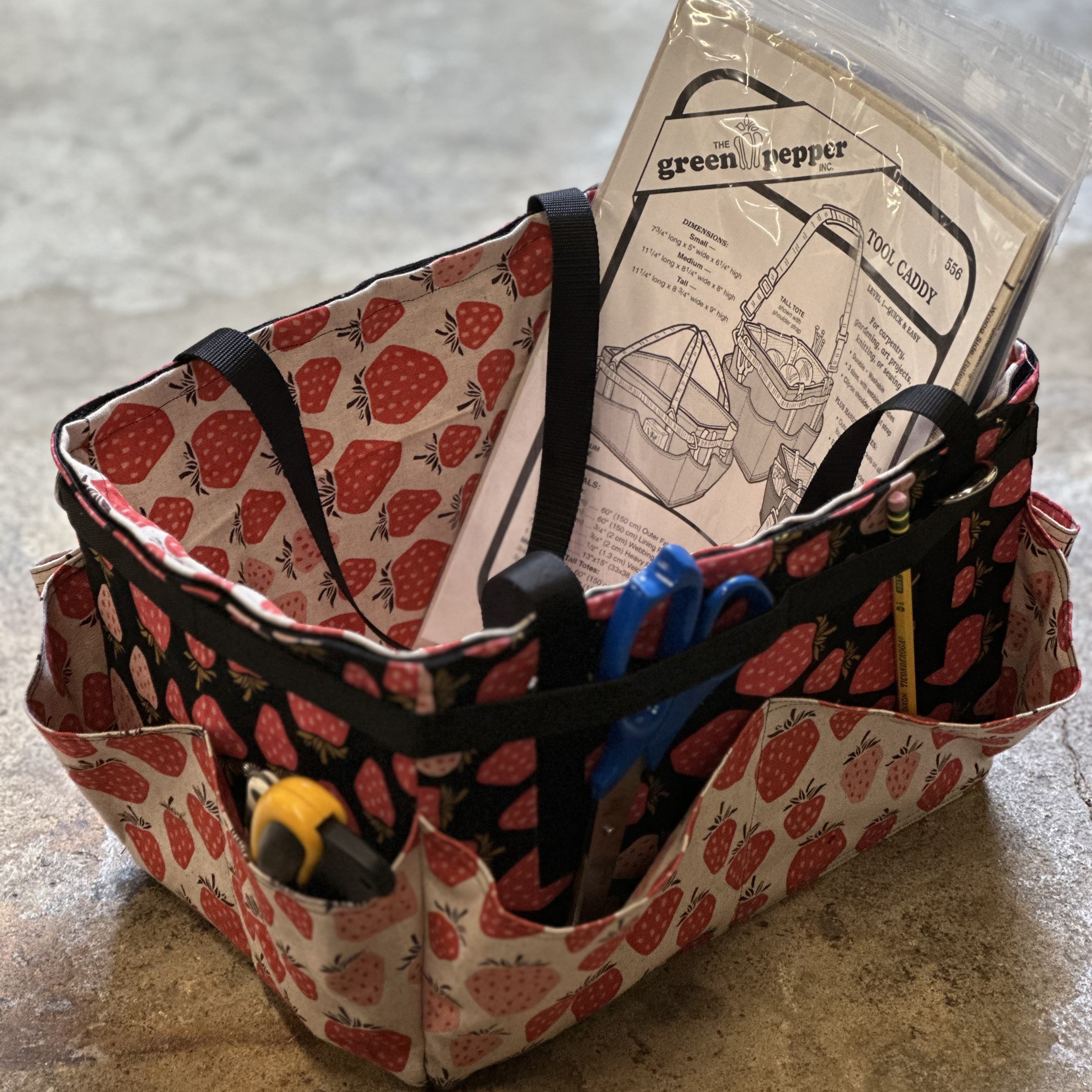 Fabric basket with strawberry pattern containing tools and a paper on a concrete surface