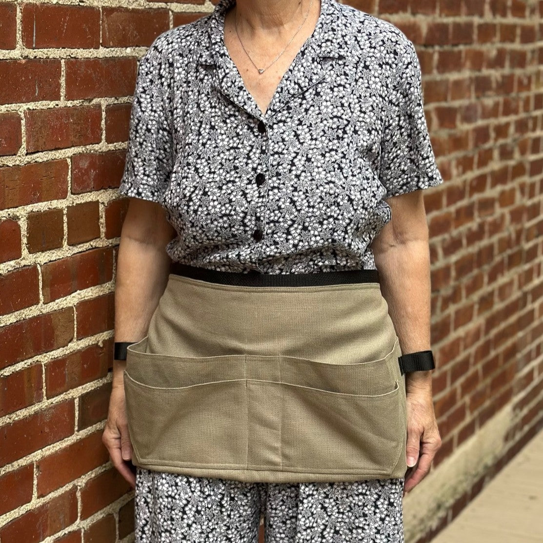 Person wearing a floral shirt and beige tool belt apron against a brick wall.