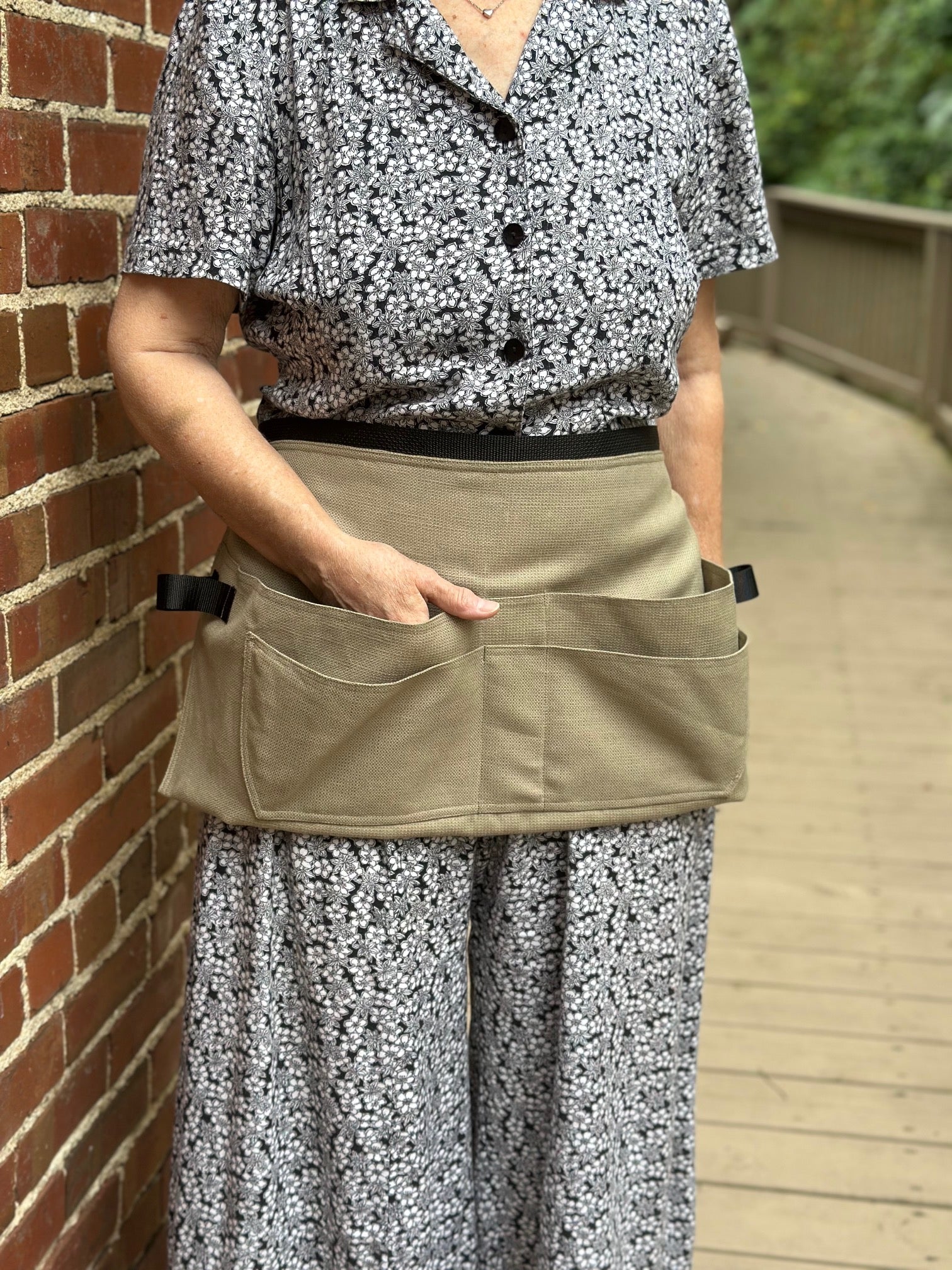 Person wearing a floral shirt and patterned pants with a beige toll belt against a brick wall.